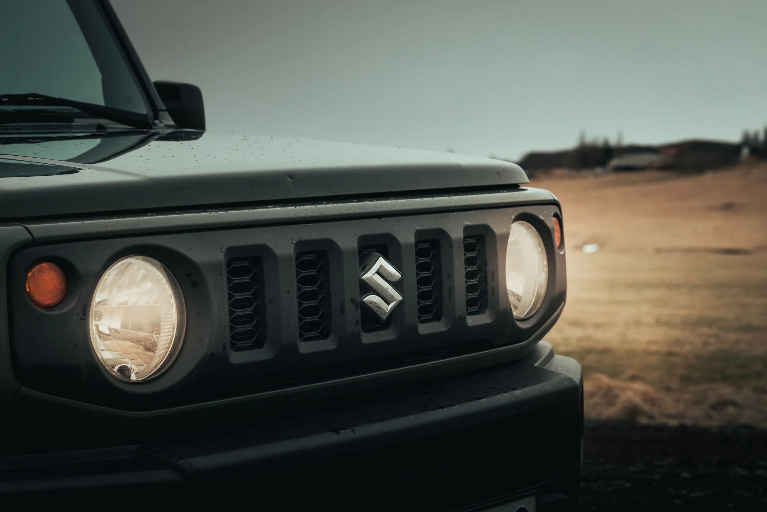 Close-up of the front of a Suzuki vehicle, showing the headlight, grille with Suzuki emblem, and part of the hood against an outdoor background.
