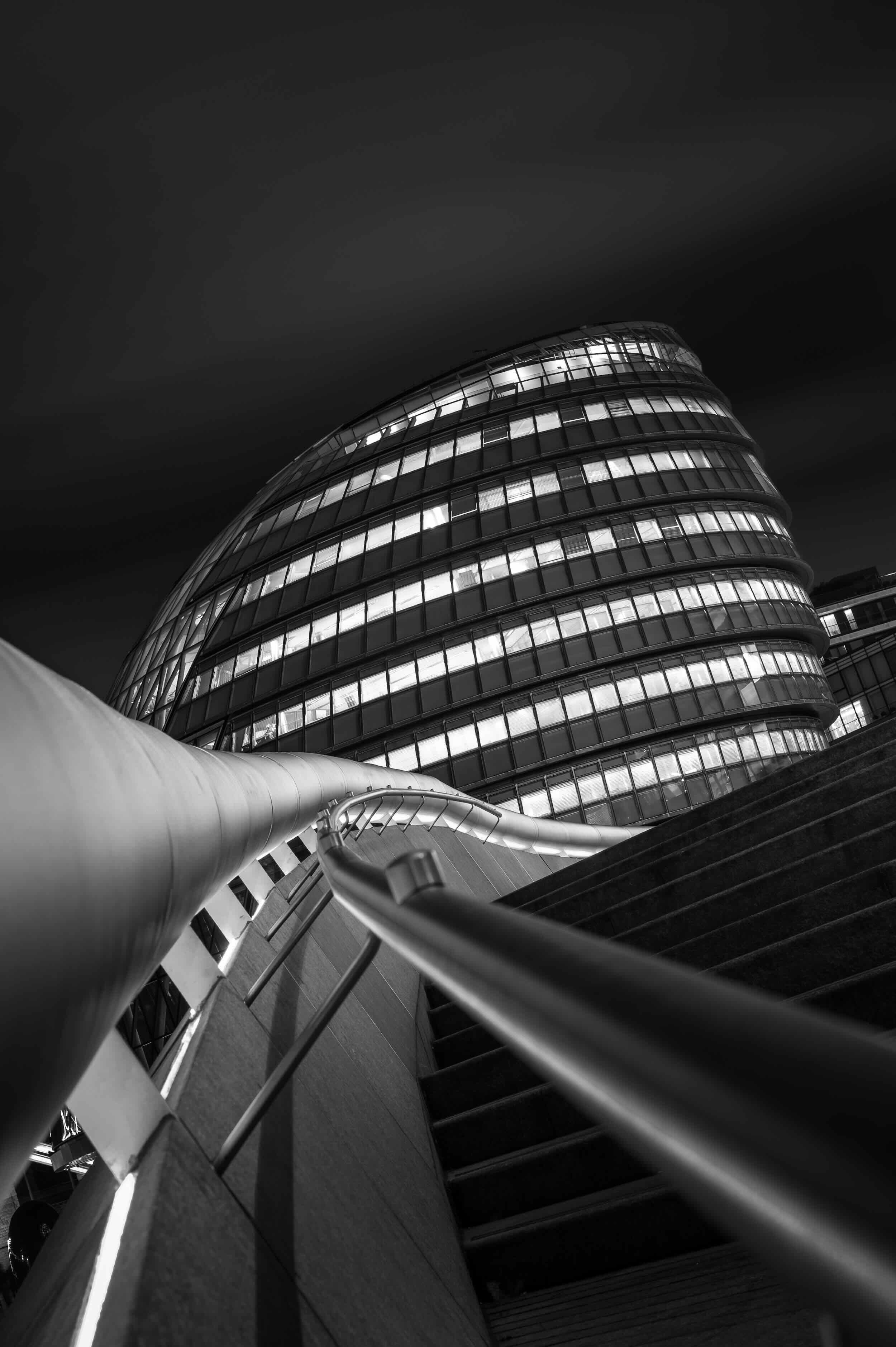 Nighttime view of a modern, curved glass building with illuminated windows, seen from a staircase with a metal handrail in the foreground.