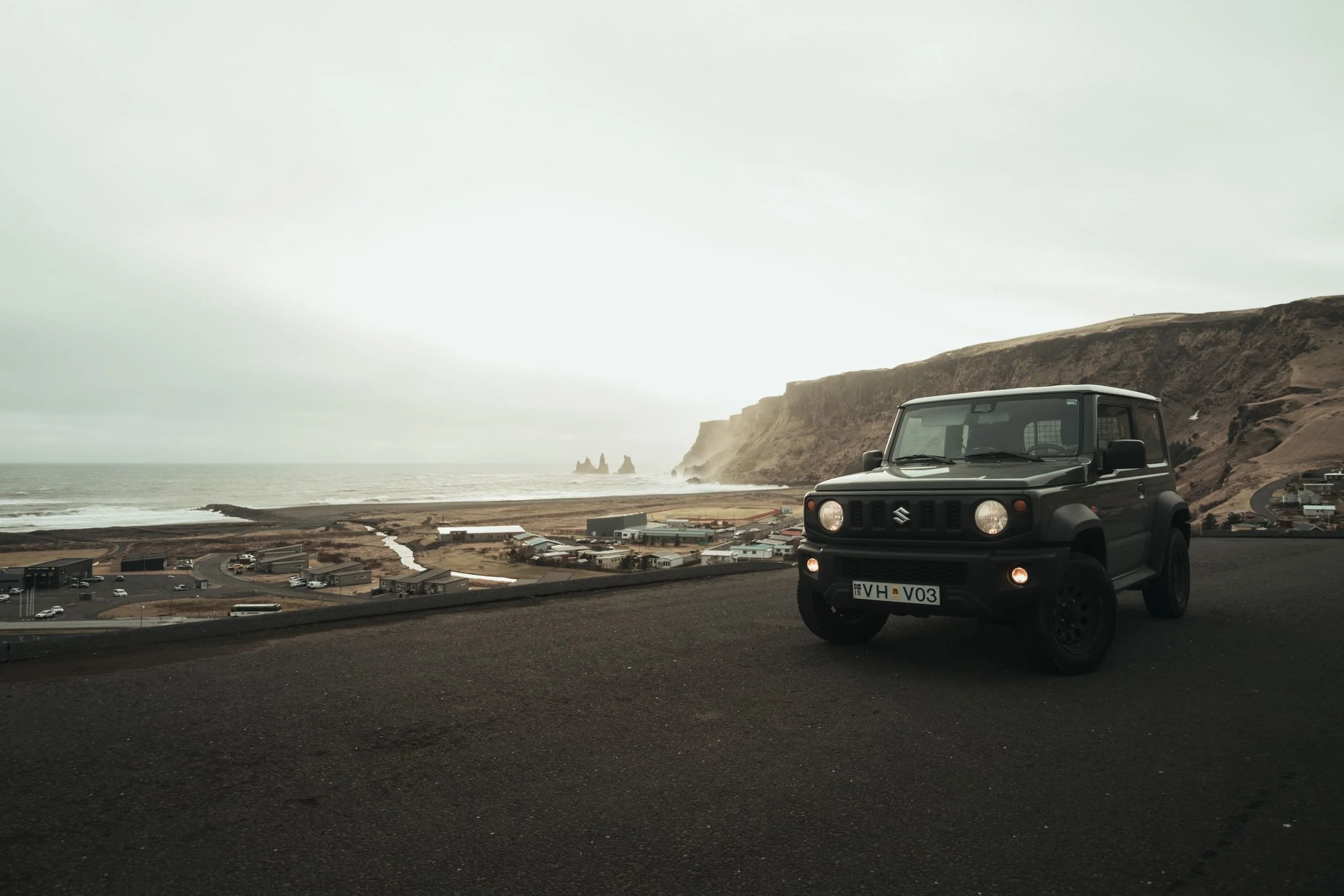 A black Suzuki Jimny parked on a hill overlooking a coastal town with cliffs and rock formations in the distance, under cloudy skies.
