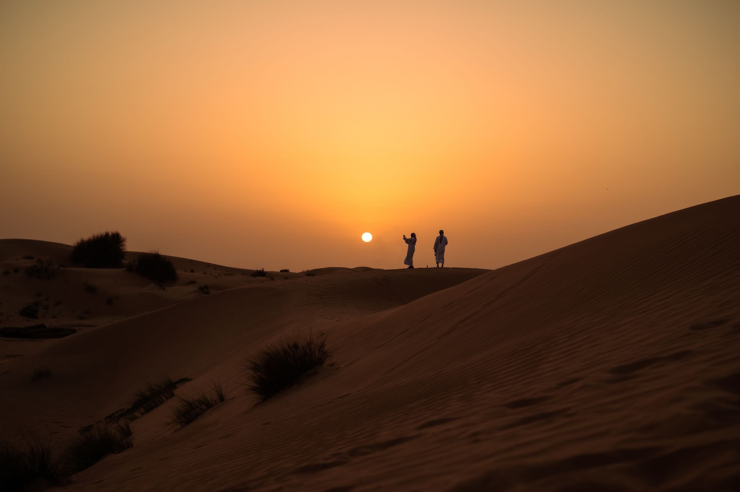 Two people in traditional Middle Eastern attire standing in a desert landscape at sunset. One is holding a device, possibly a phone or camera, while the other stands nearby. Sand dunes and sparse vegetation are visible, with a large setting sun on th