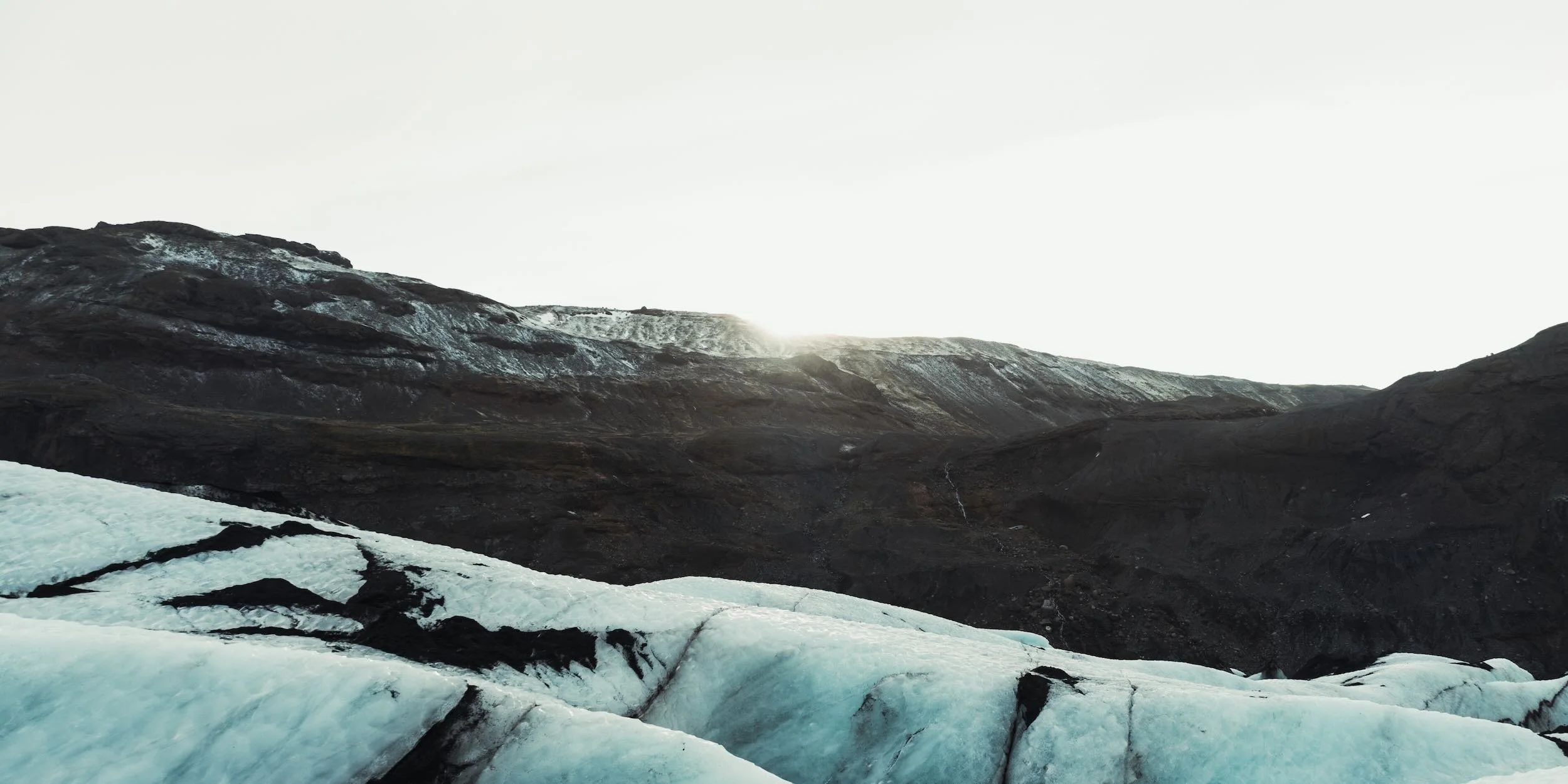 Glacier in a mountainous landscape with a bright sky, dark rocky terrain, and patches of ice and snow.