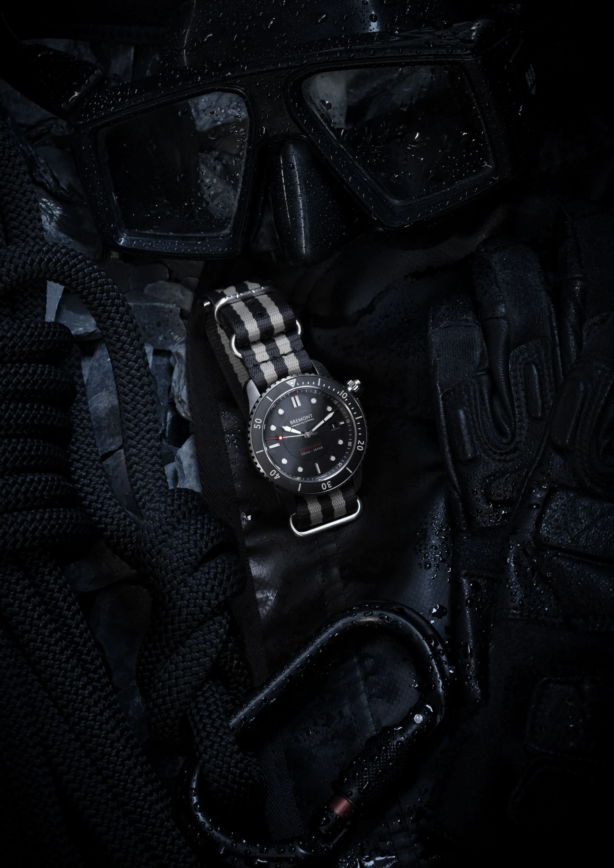 A water-resistant wristwatch with a black face and striped fabric strap resting on wet gear, including a pair of goggles with water droplets and black gloves, surrounded by water droplets.