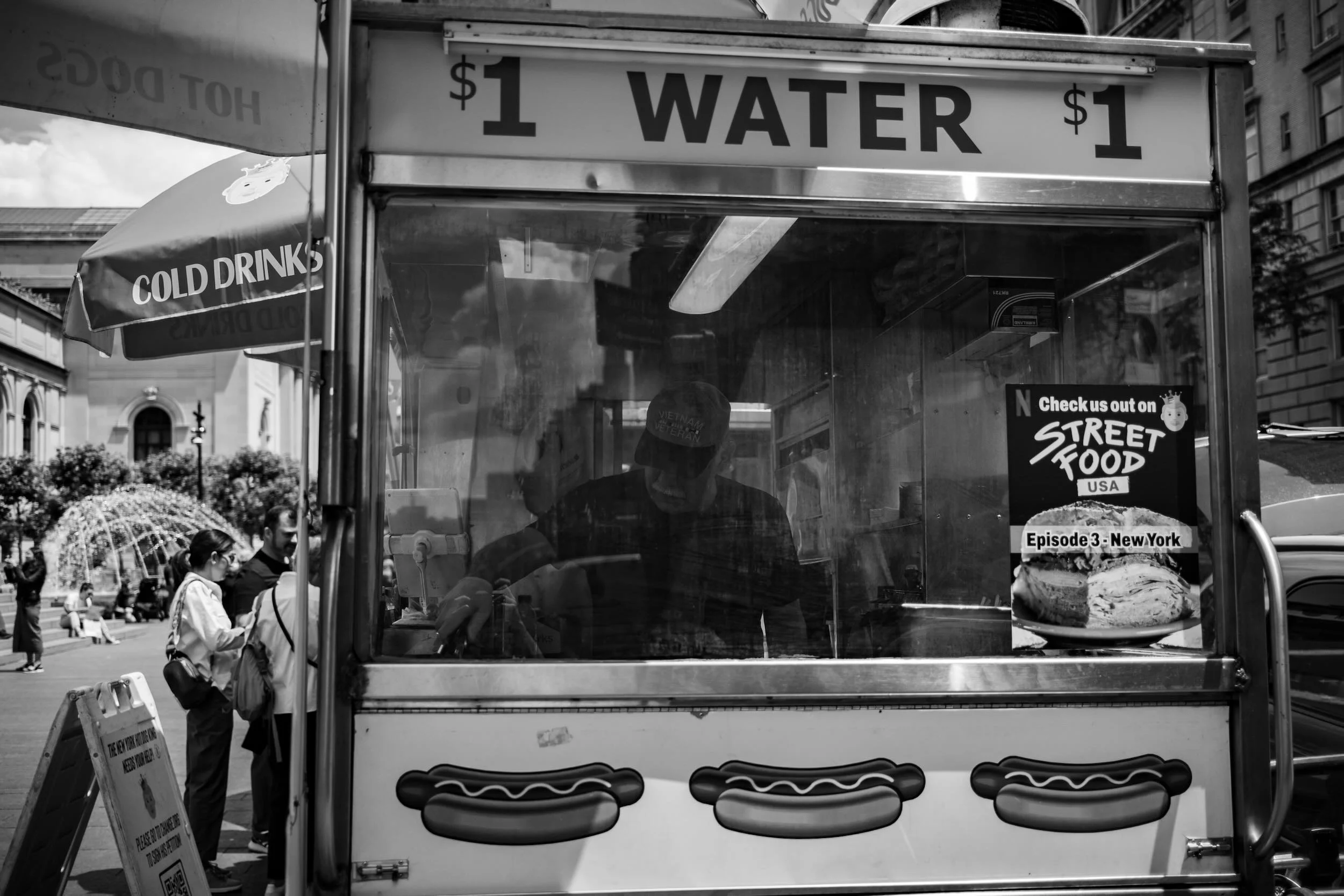 A street vendor stand labeled 'Water $1' with a sandwich advertisement, in an urban setting with pedestrians and historic buildings in the background, black and white photo.
