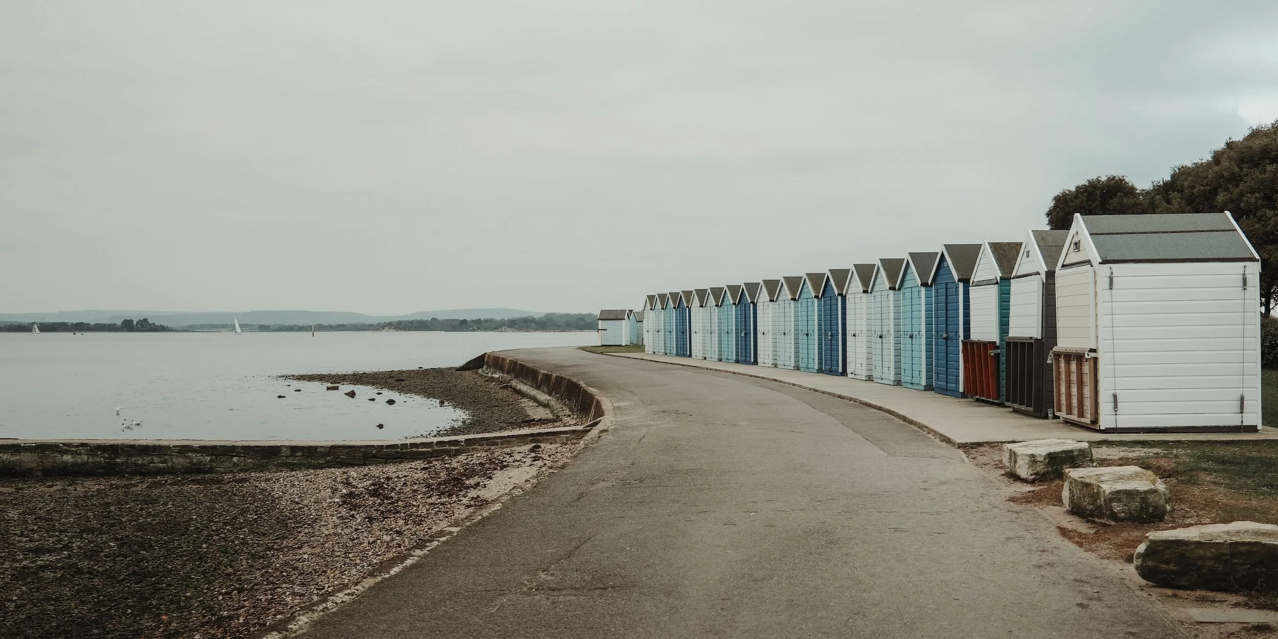 A row of colorful beach huts along a lakeside promenade with sailboats in the background under an overcast sky.