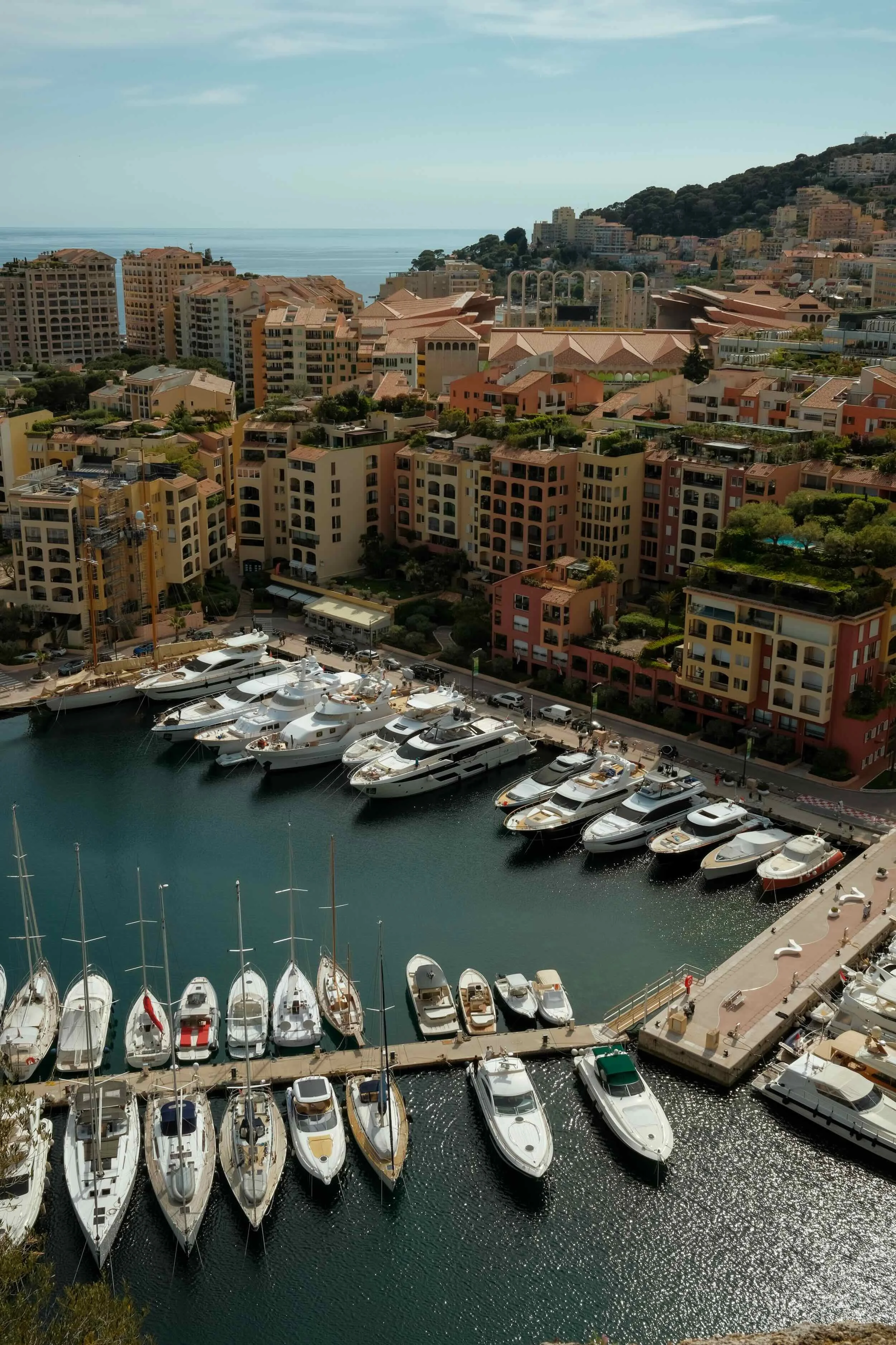 A marina filled with luxury yachts docked along a harbor, with a cityscape of colorful Mediterranean-style buildings and hills in the background, and the ocean visible in the distance.