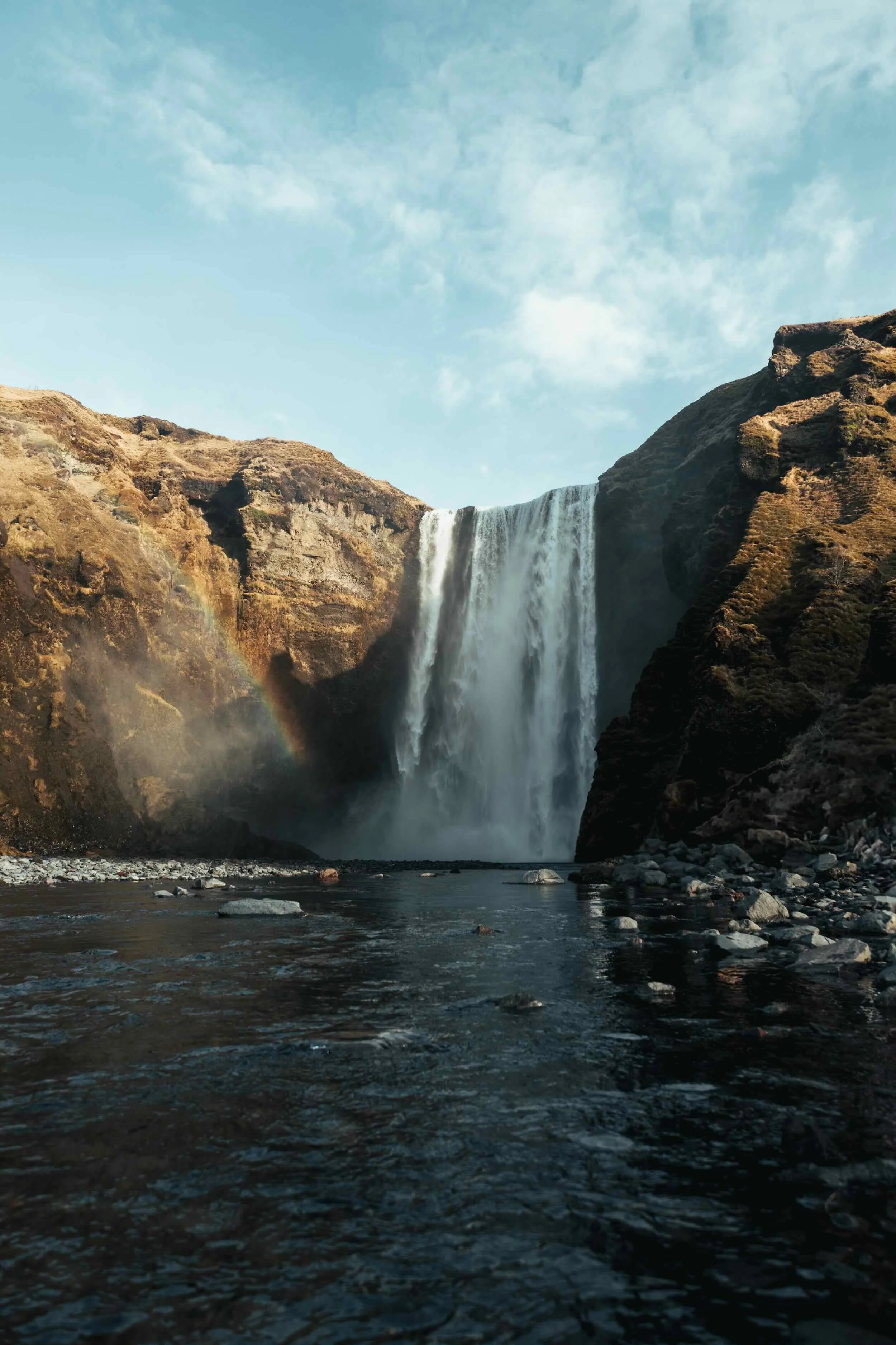 A tall waterfall flowing between rocky cliffs with a rainbow visible near the base, set against a partly cloudy sky.
