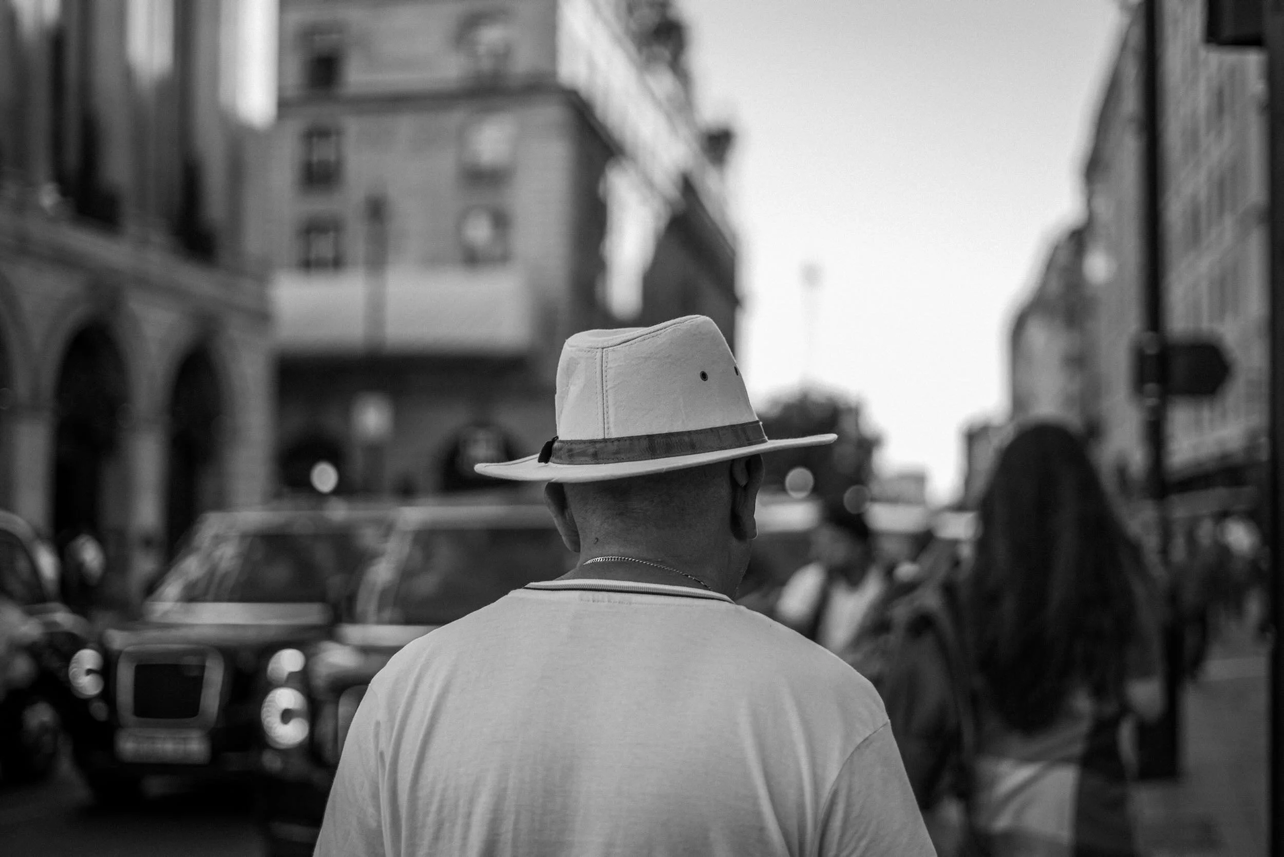 A person wearing a broad-brimmed hat and a T-shirt, seen from behind, standing on a city street with buildings and cars in the background.