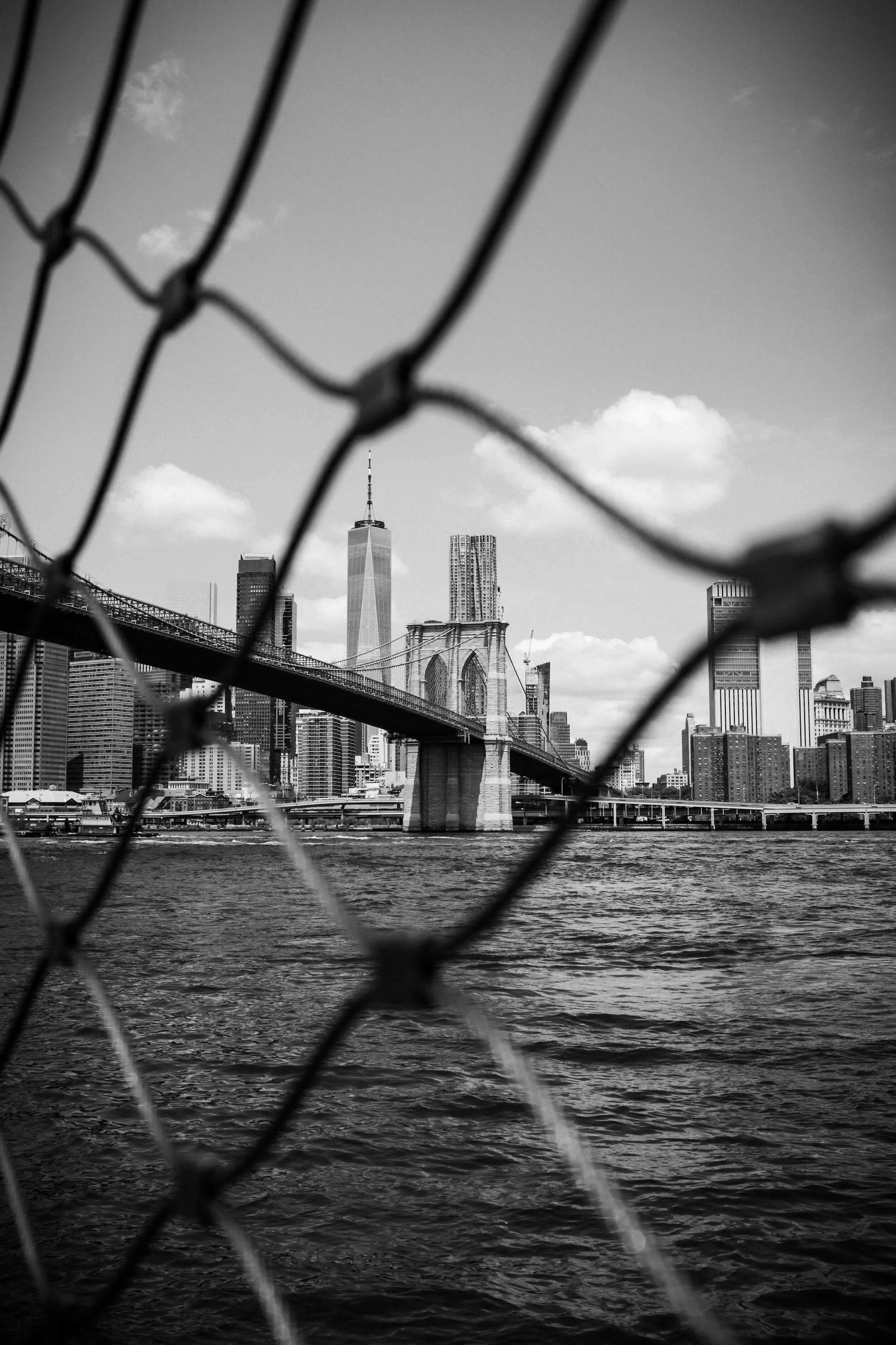 Black and white photograph of a city skyline with tall buildings, including one with a spire, seen through a chain-link fence over a body of water, with the Brooklyn Bridge visible in the foreground.