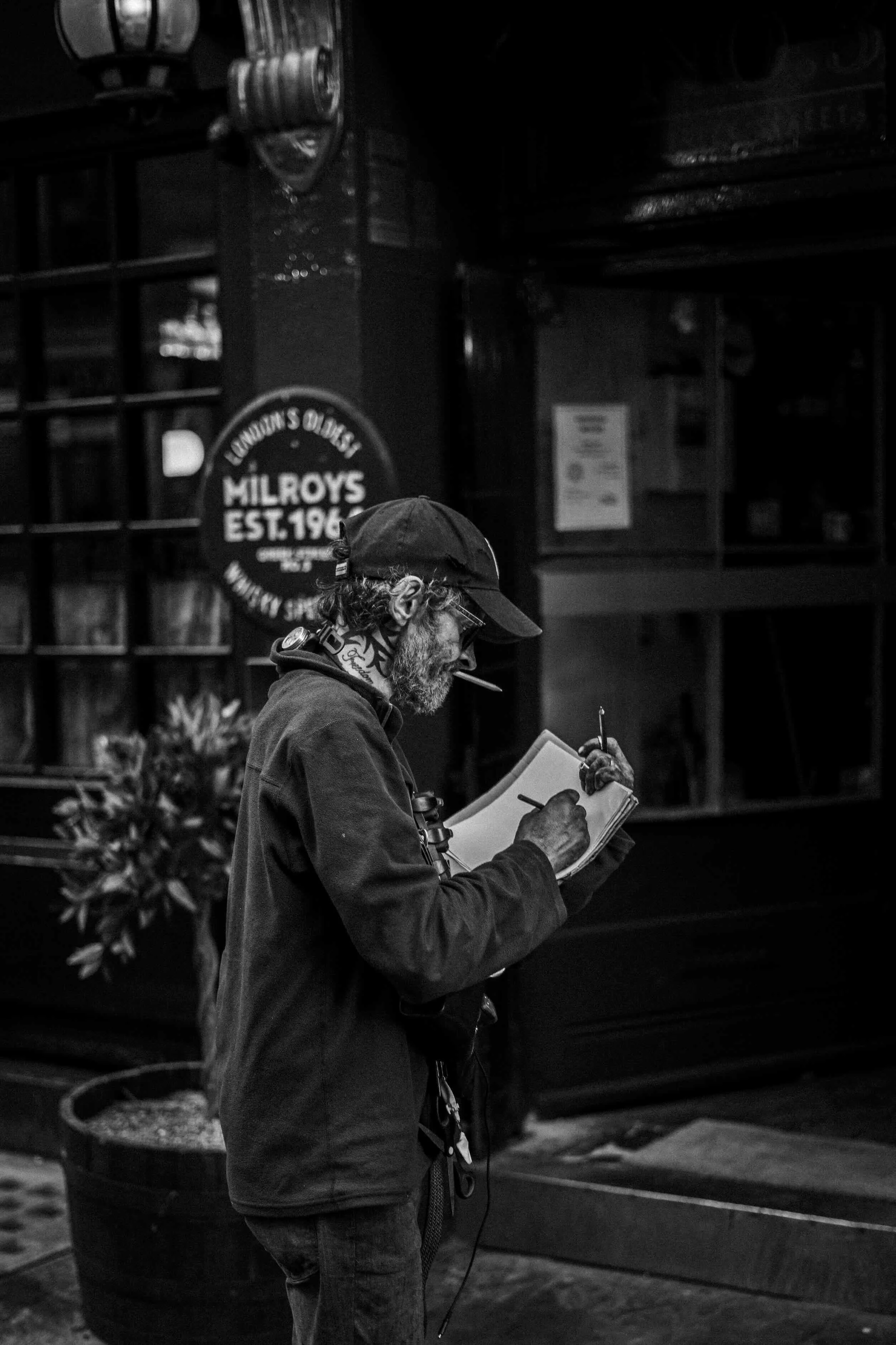 A man with curly hair, glasses, and a beard is standing outside in front of a wall with signs, holding a notebook and a pen, wearing headphones. The image is in black and white.
