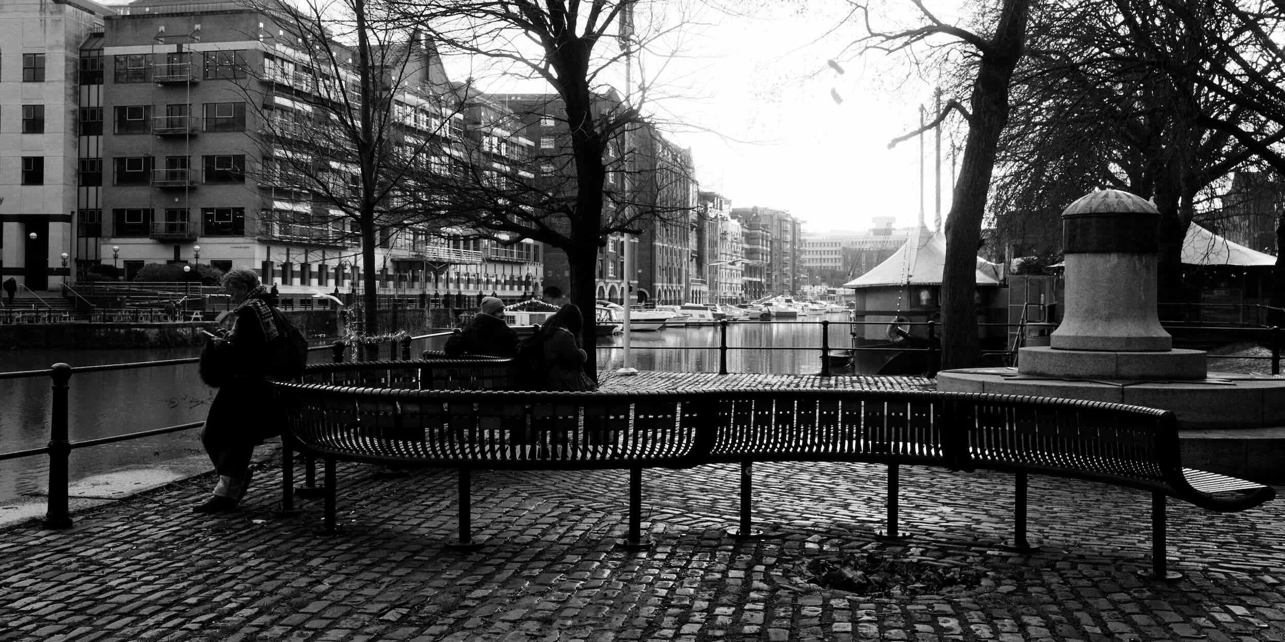 A black-and-white photo of a riverside scene with three people sitting and standing on a cobblestone path, leafless trees, modern buildings across the water, boats docked along the shore, and a gazebo with string lights.