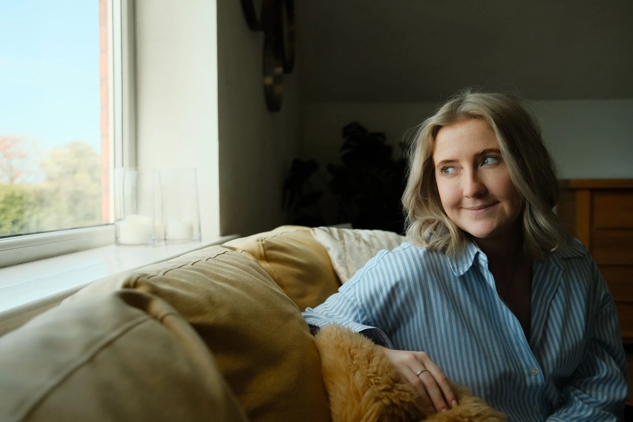 A woman with blonde hair and wearing a striped shirt sitting on a beige couch near a window, looking at a golden retriever dog that is lying on the couch with its paws on her lap.