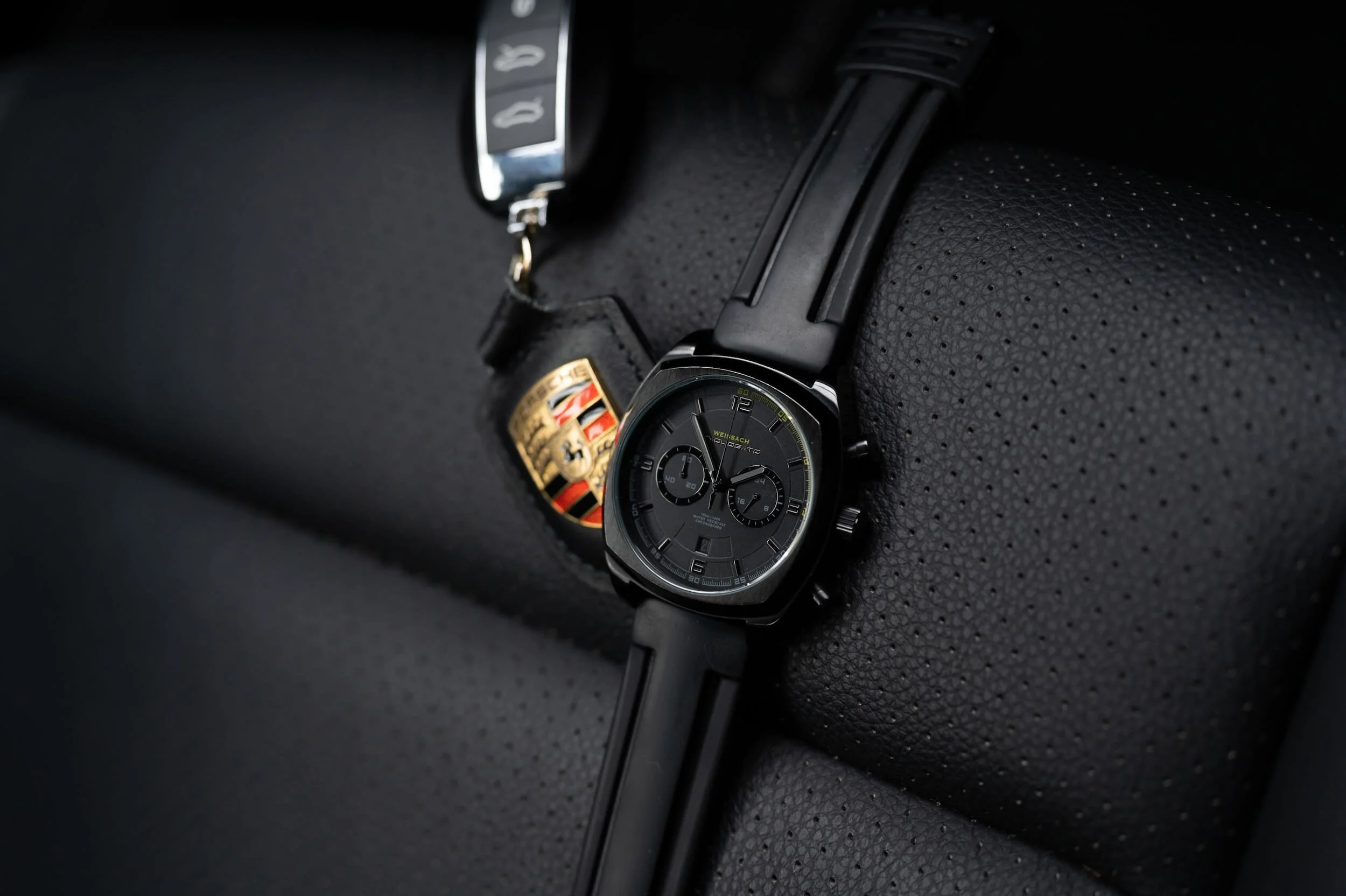 A luxury Porsche watch with a black strap placed on a black textured surface, alongside a Porsche keychain.