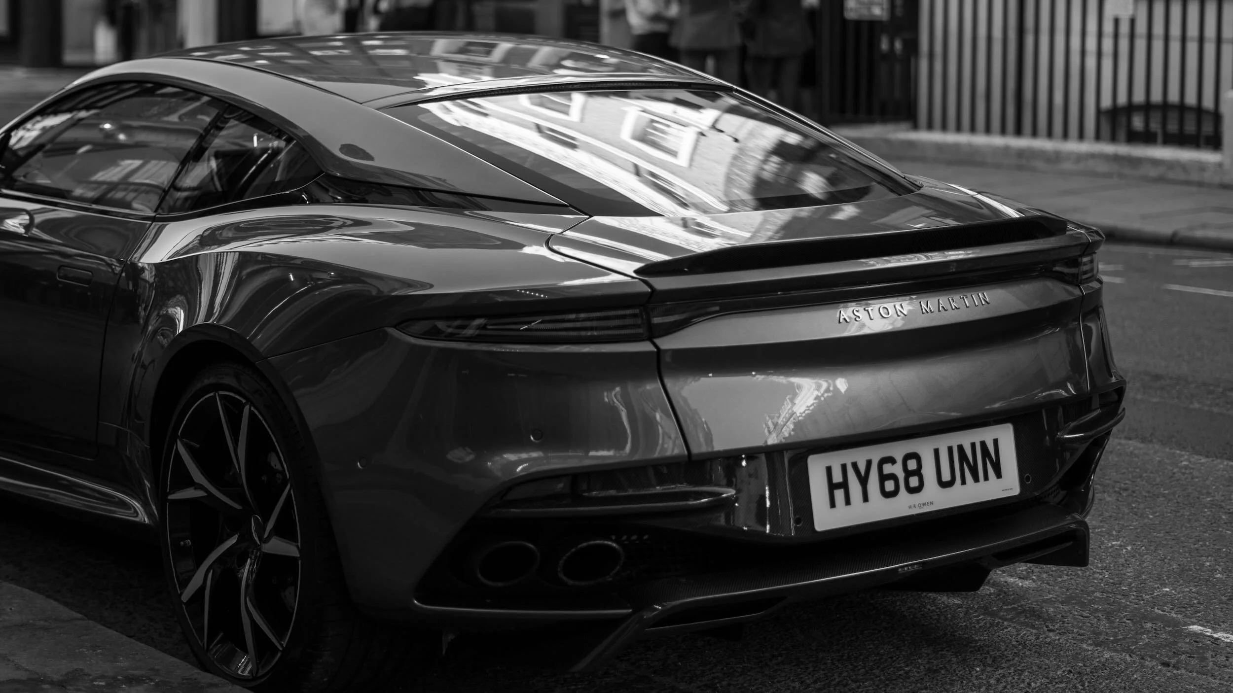 A sleek, modern Aston Martin sports car parked on a city street, shown from the back and side in black and white.