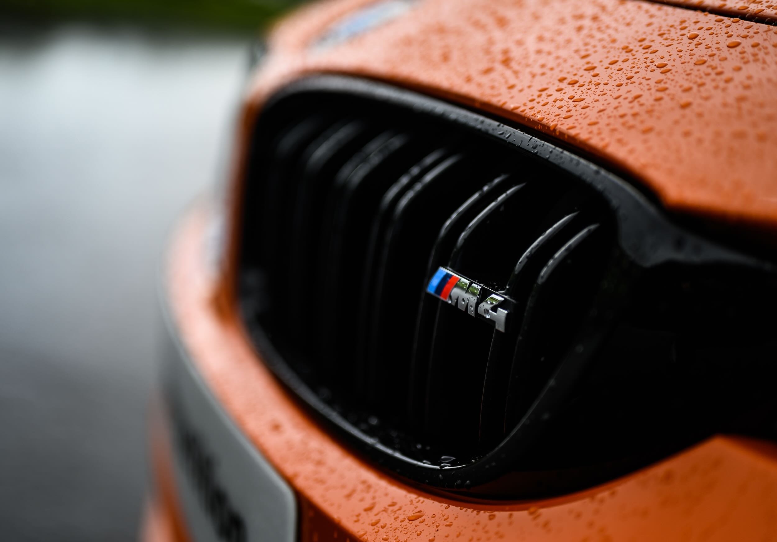 Close-up of the front grille of a car with an orange hood, rain droplets on the surface, and a BMW M logo.