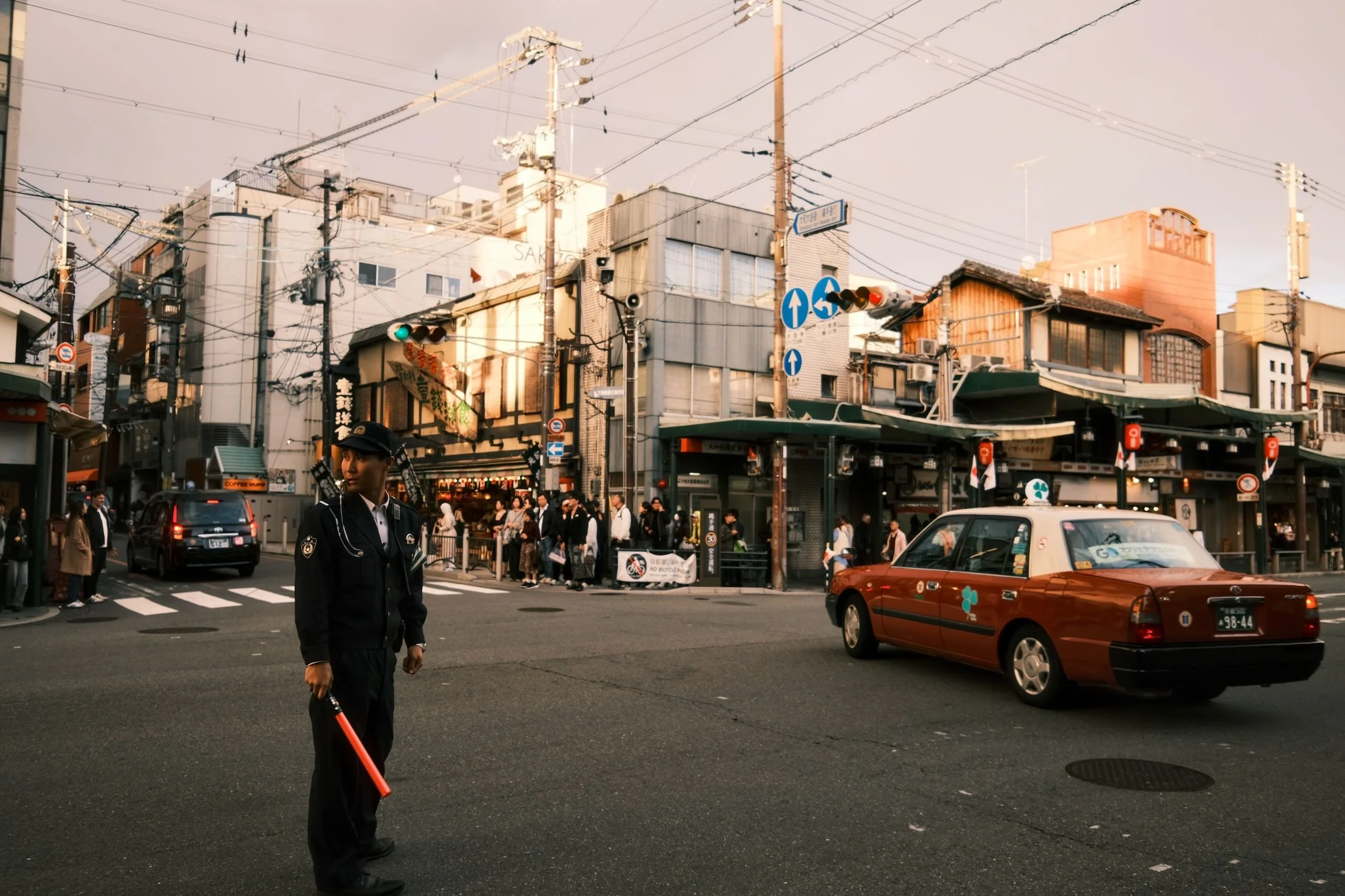 A busy urban street scene with pedestrians, cars, and buildings. A police officer stands in the foreground, with power lines overhead and a crowd near storefronts in the background.