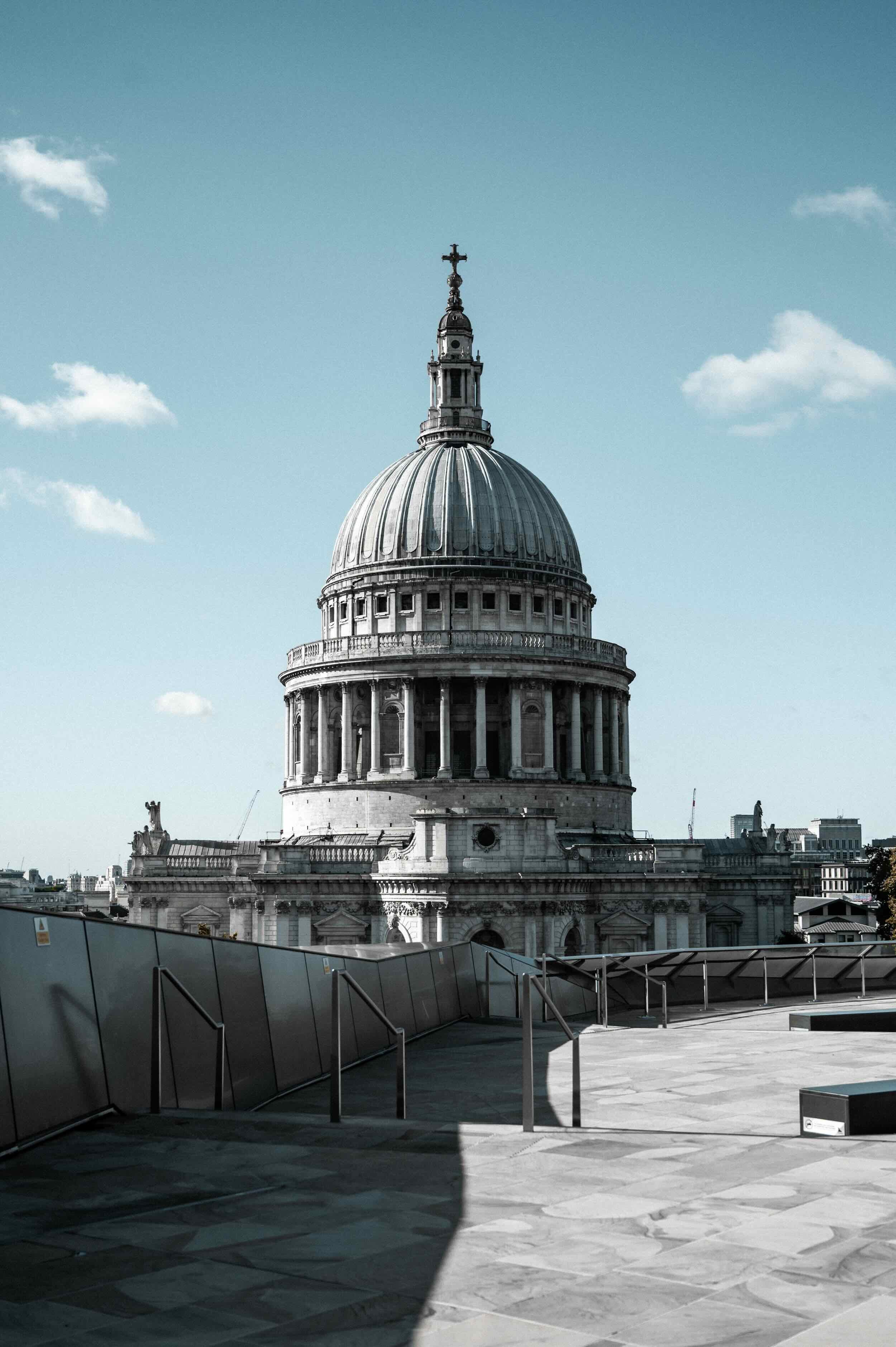 The image shows the iconic dome of St. Paul's Cathedral in London, England, with a clear blue sky and some clouds in the background. The foreground includes a modern rooftop terrace with glass panels and metal railings.