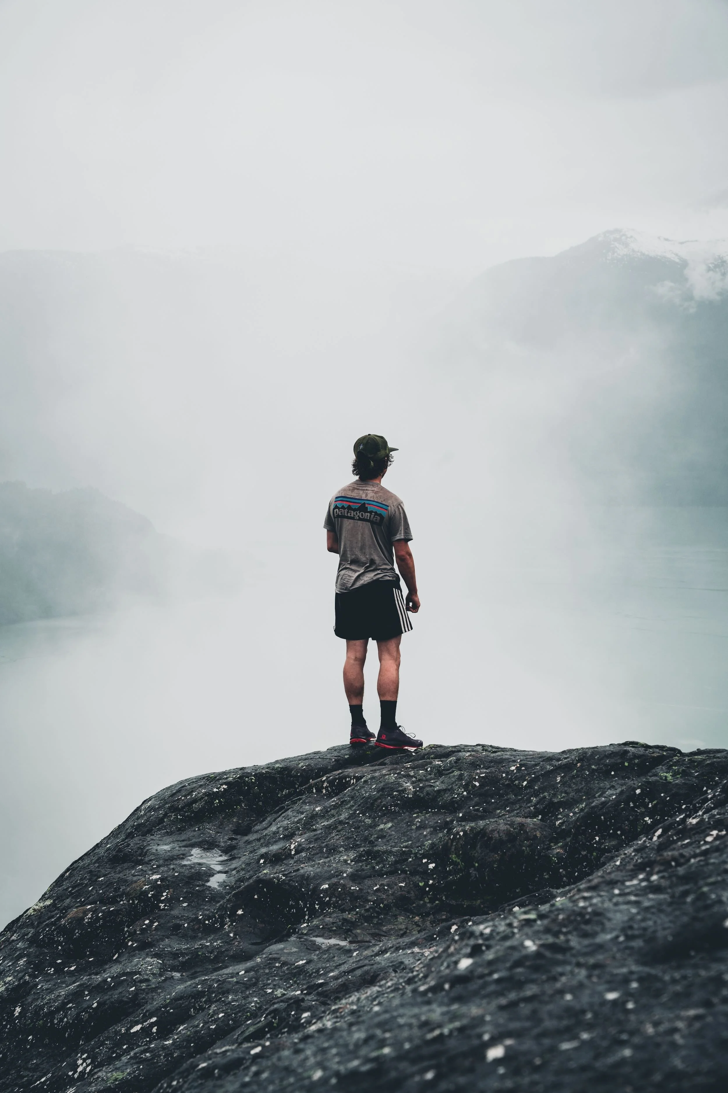 A person standing on a large rock, looking out at a foggy mountainous landscape. The person is wearing a Patagonia t-shirt, shorts, and hiking shoes, with their back to the camera.