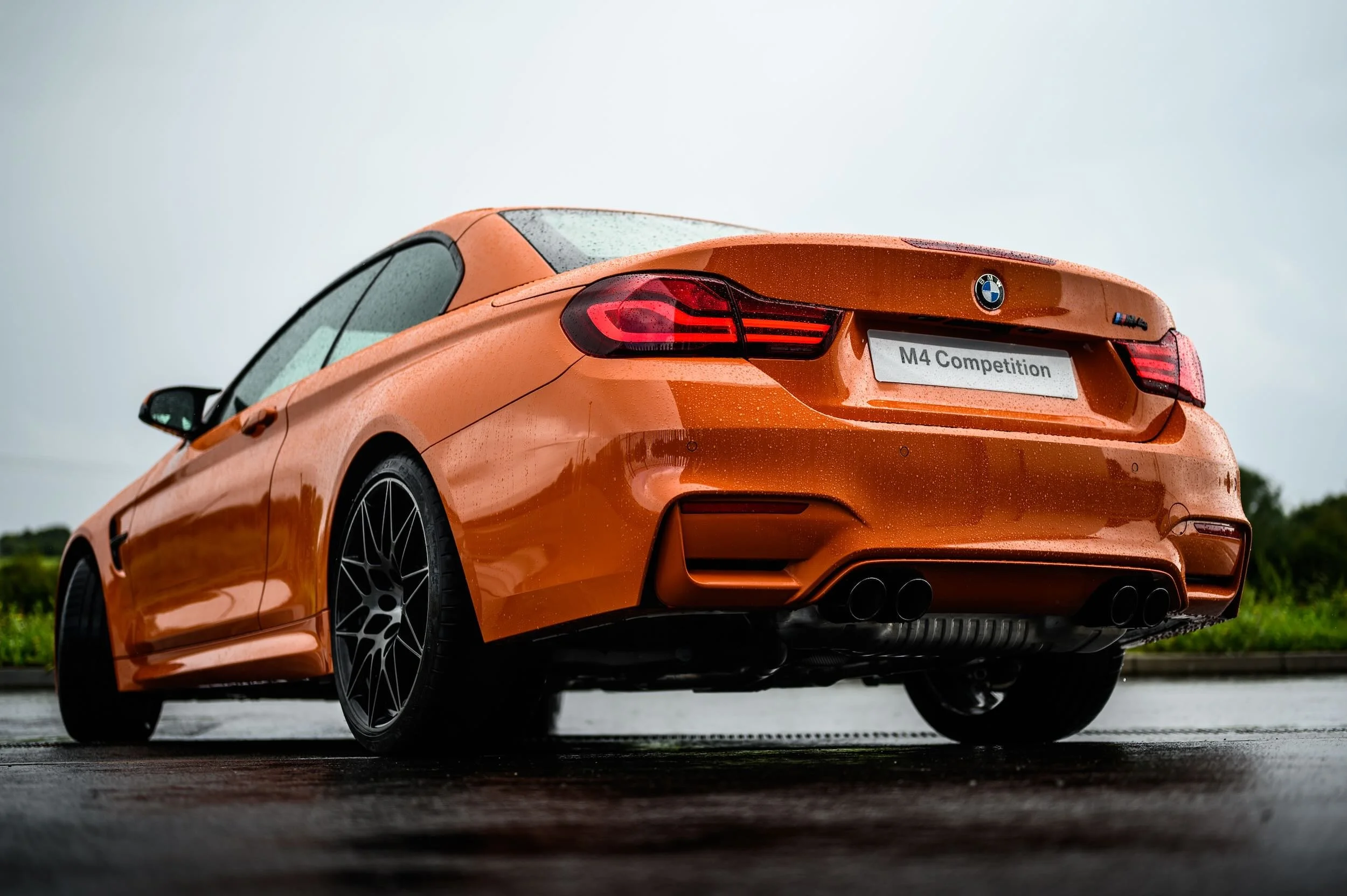 An orange BMW M4 Competition sports car parked on a wet road during overcast weather, with raindrops visible on the car's surface.