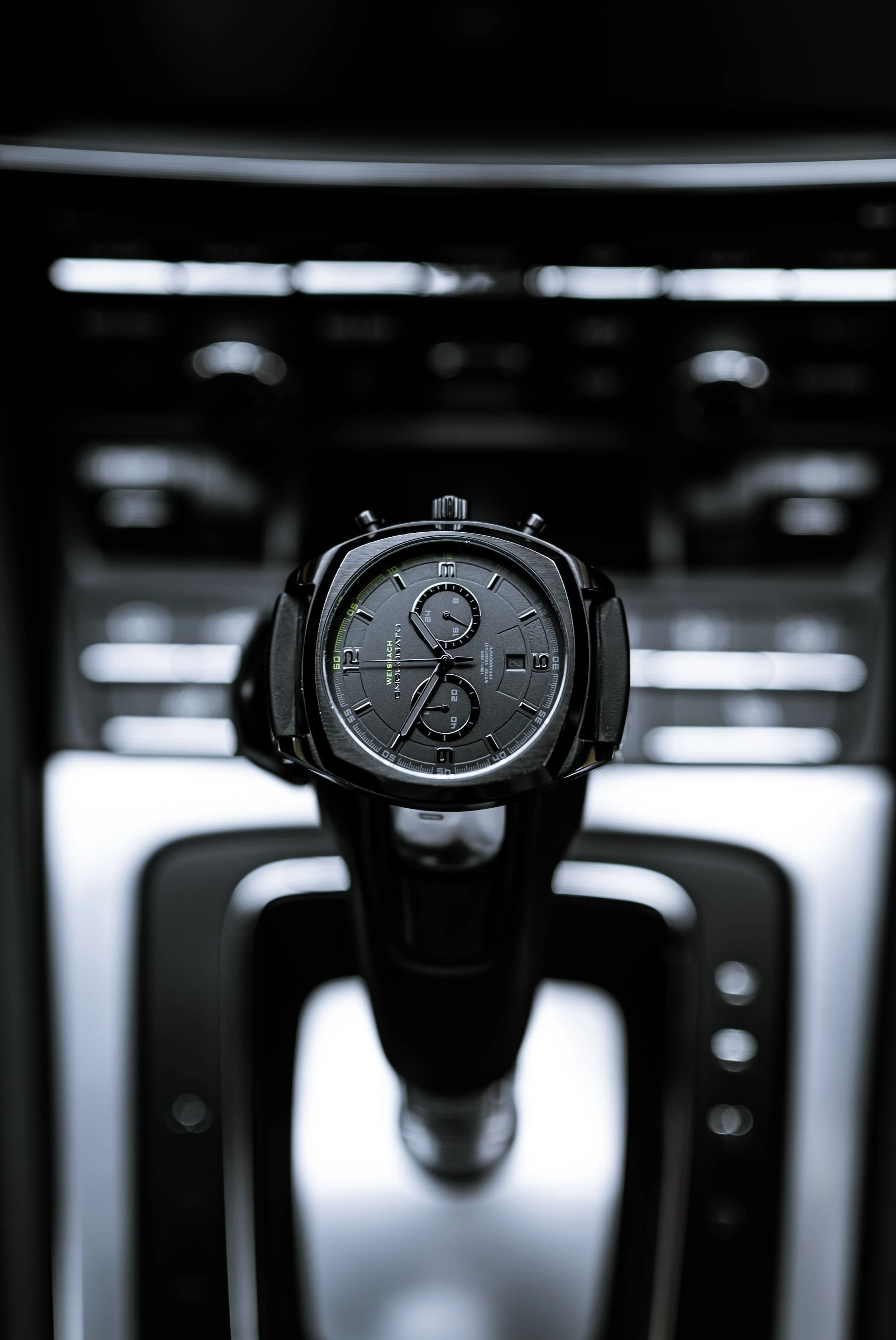Close-up of a black wristwatch with multiple dials, mounted on a metallic gear shift in a vehicle interior.