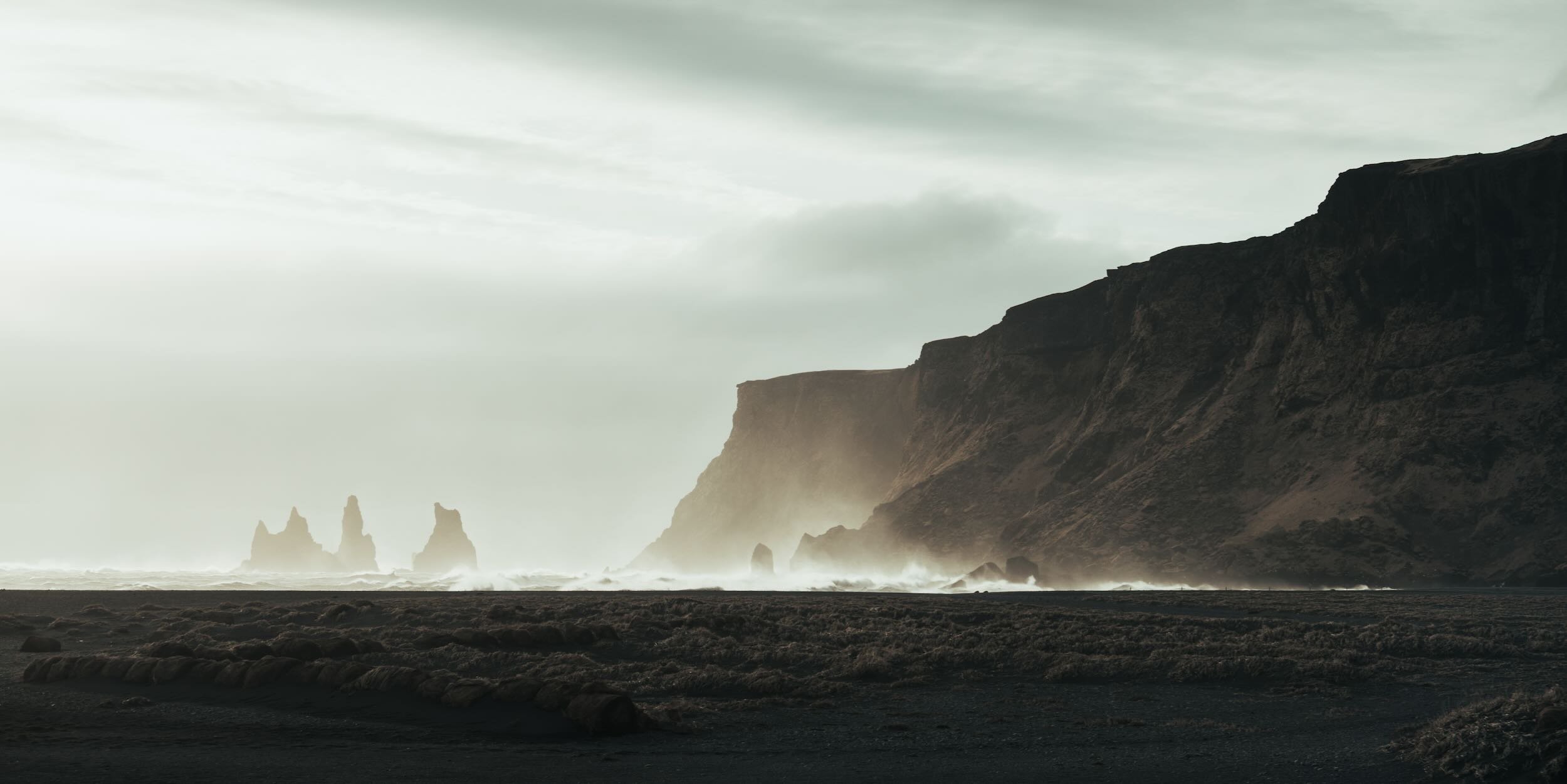 A rugged coastal landscape with large cliffs on the right side, sea stacks in the distance, and waves crashing against the shore under a cloudy sky.