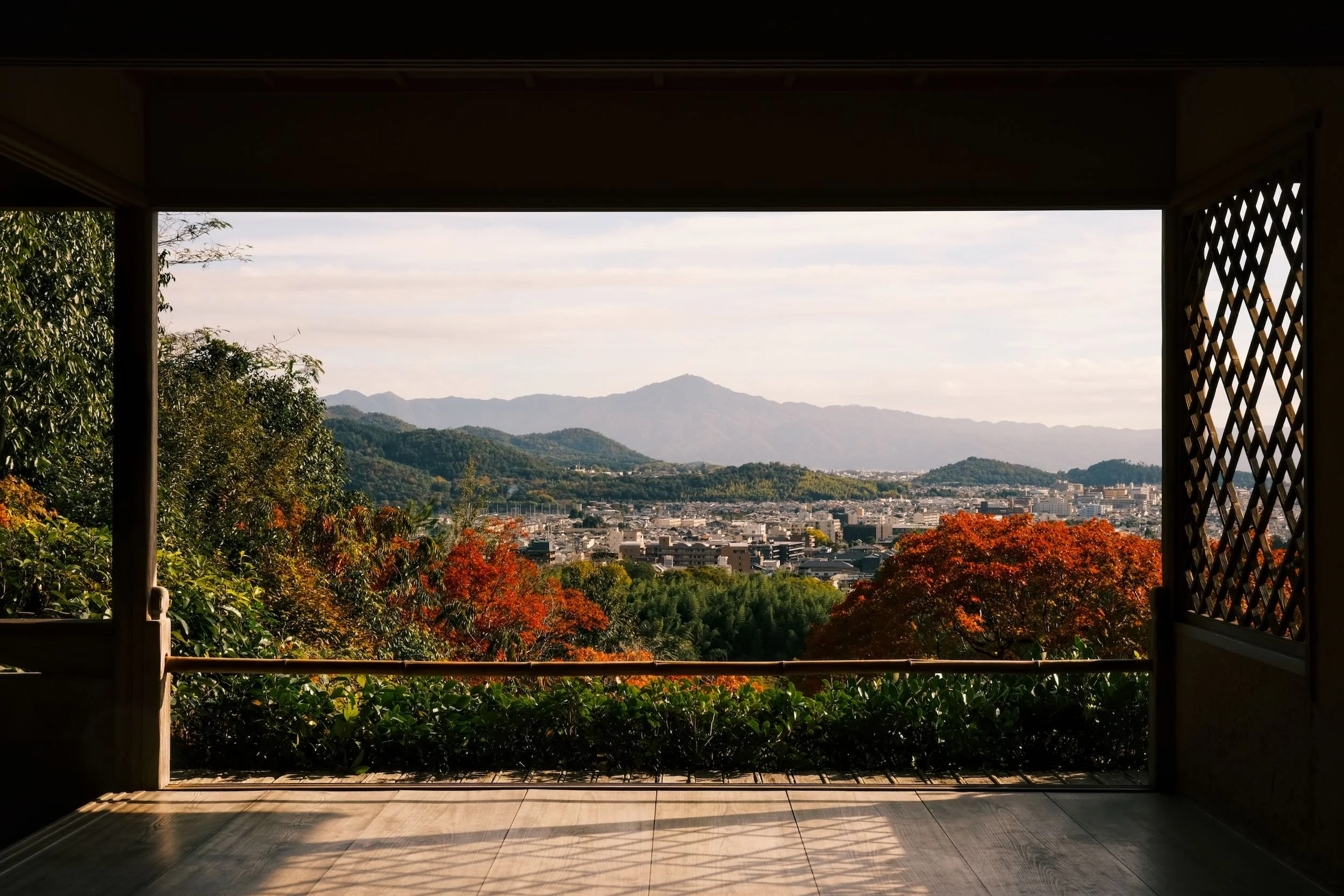 View from a traditional wooden porch overlooking a cityscape with mountains in the background, trees with autumn foliage, and a partly cloudy sky.