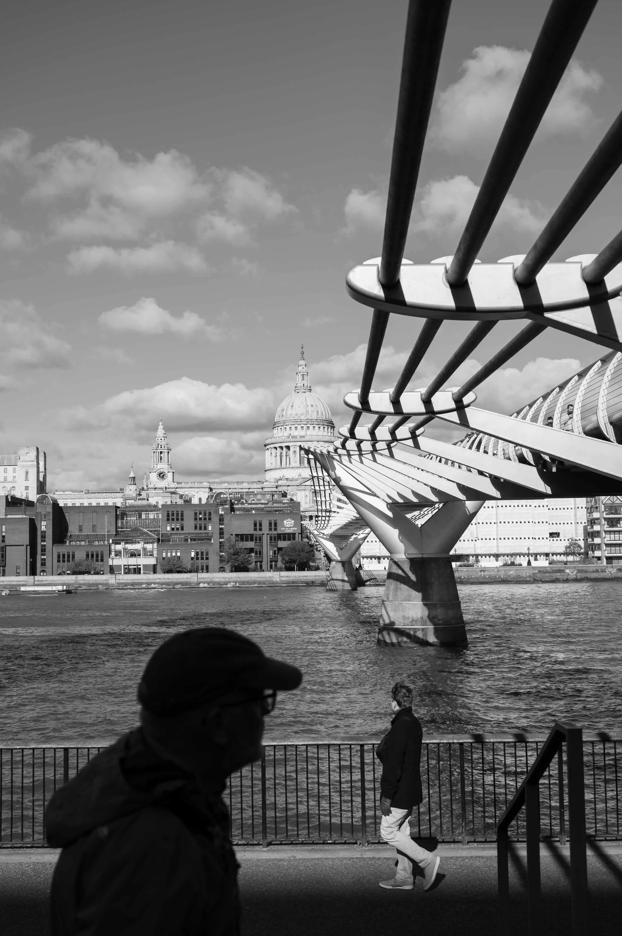Black and white photo of people walking along a riverfront promenade with the city skyline and a domed building in the background, and a modern bridge with curved supports in the foreground.