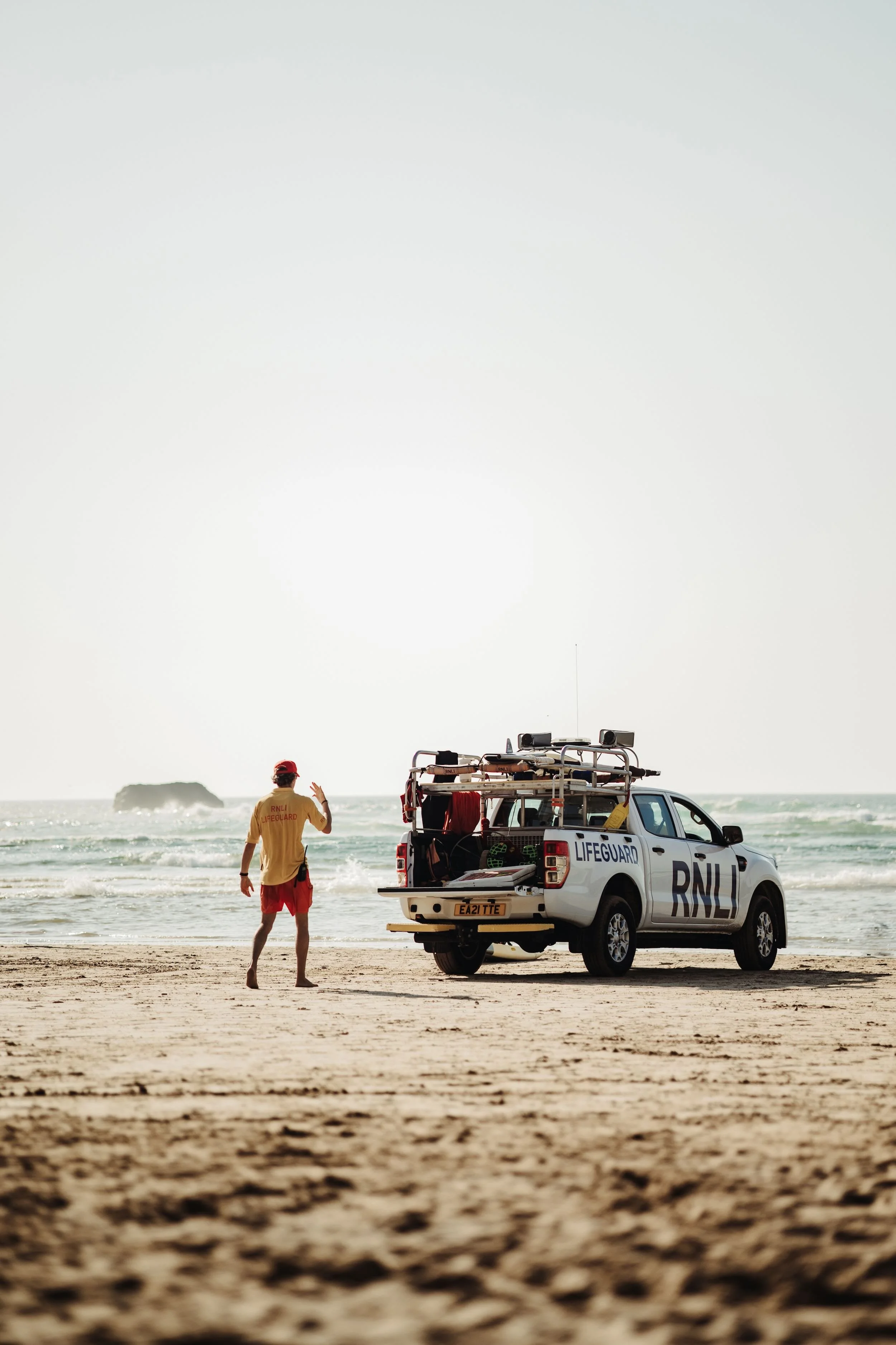 A lifeguard standing on the beach near the ocean, wearing a yellow shirt and red shorts, with a white lifeguard vehicle nearby. The vehicle has the word 'LIFEGUARD' on its side and equipment on the roof. In the background, a person in a red cap and s
