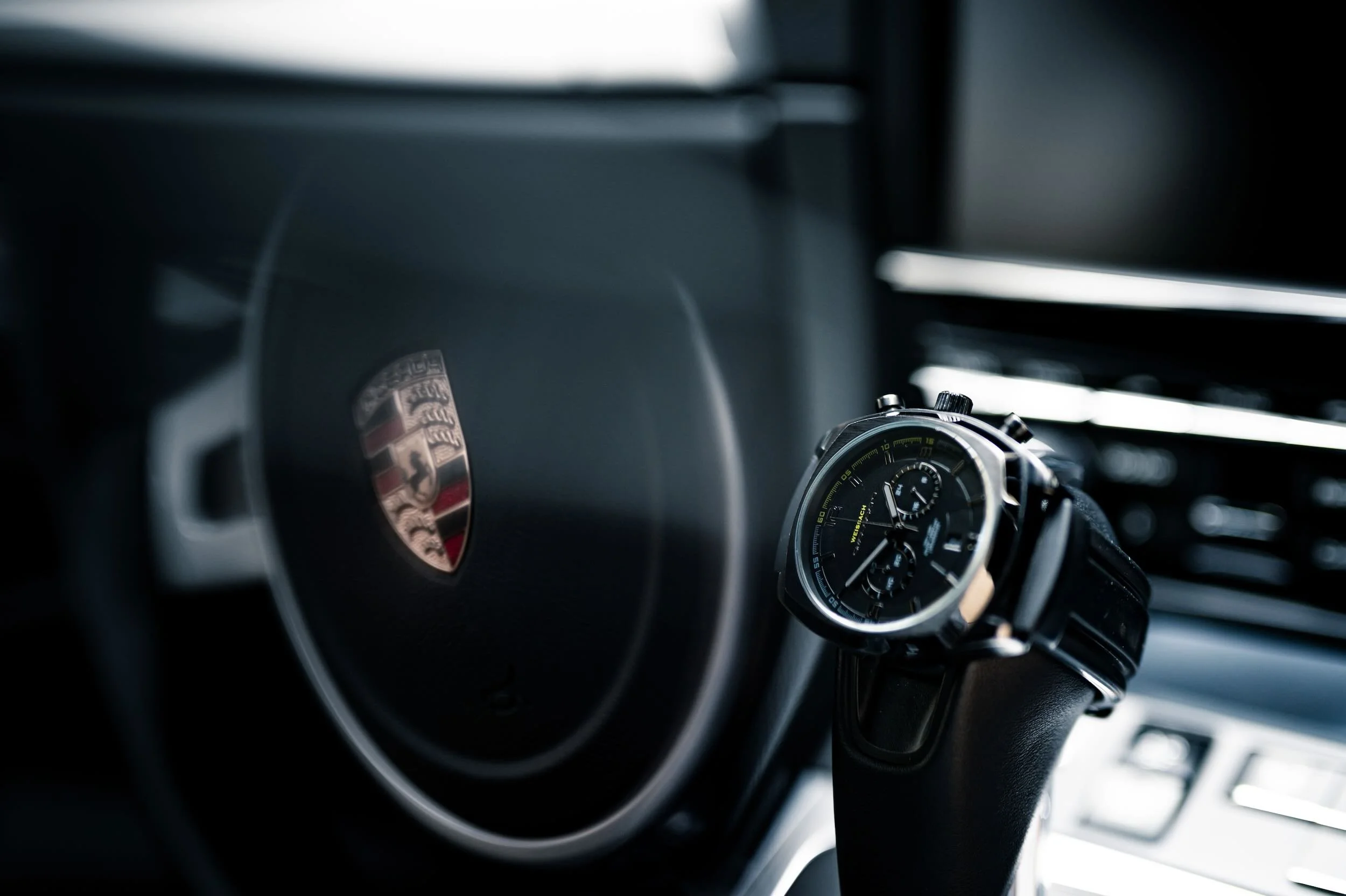 Close-up of a luxury car's dashboard and steering wheel, featuring the Porsche emblem and an analog wristwatch with a black dial and multiple subdials.