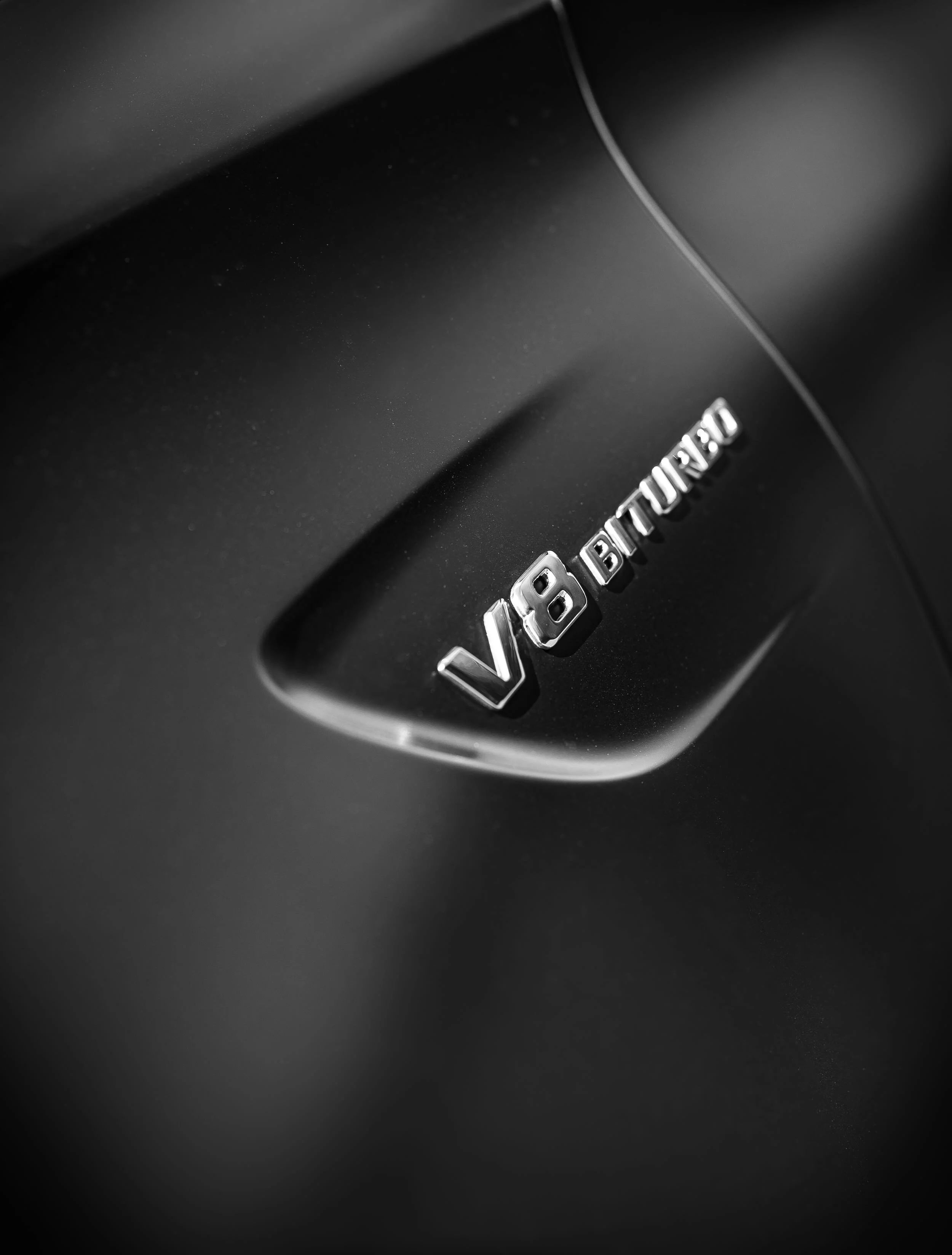 Close-up of a black car's rear with chrome letters spelling 'V8 BITURBO' on the trunk.