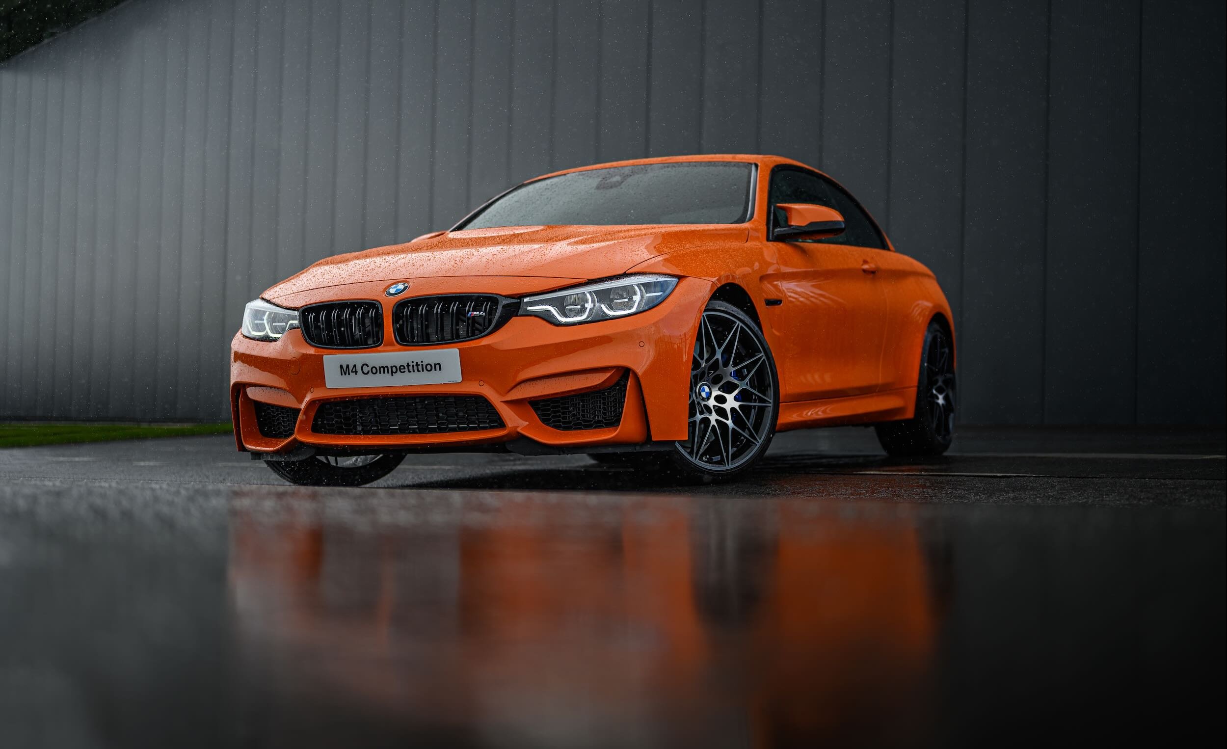 Orange BMW M4 Competition sports car parked on a wet surface with rain droplets on the car, against a dark grey building background.