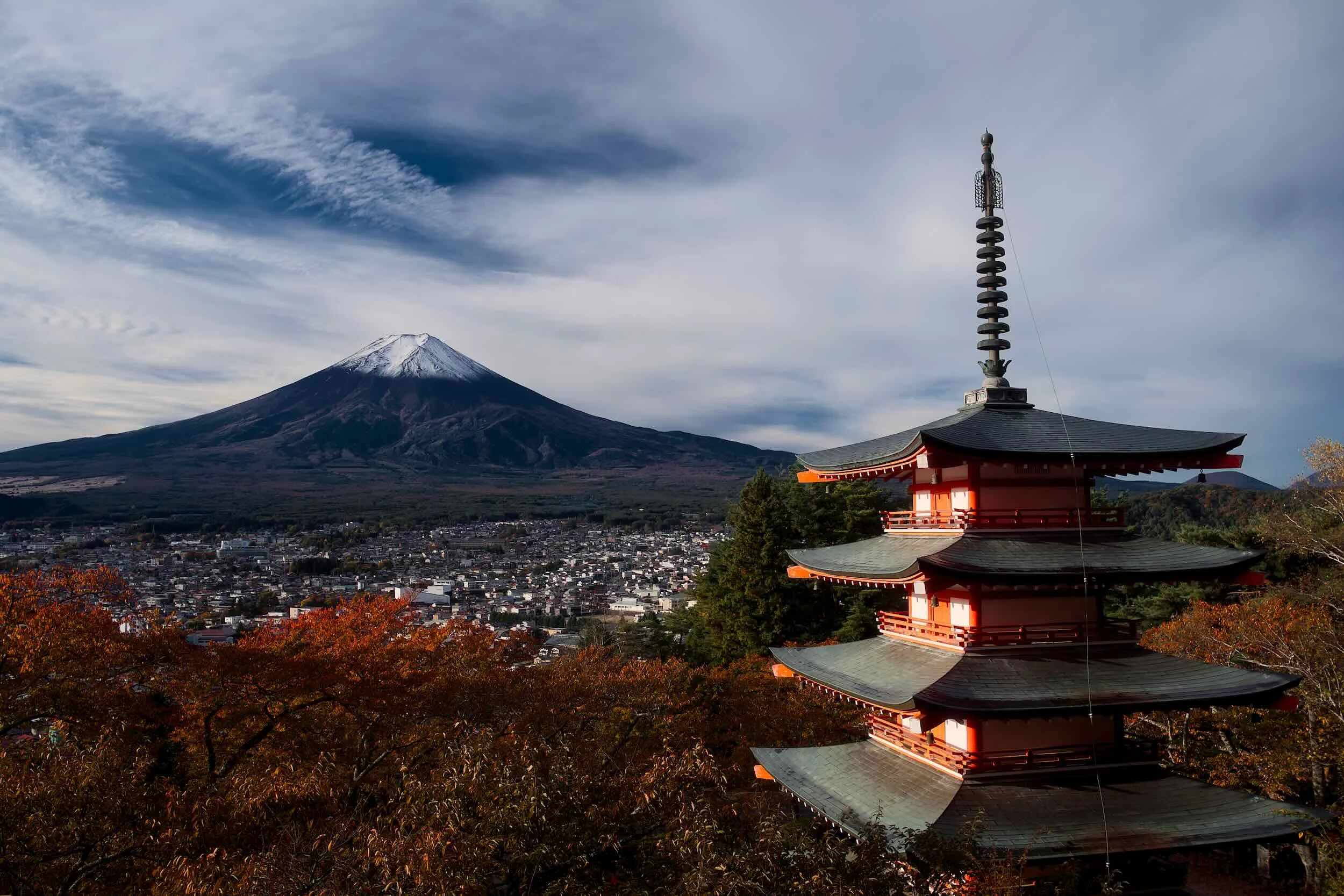 A pagoda-style building with multiple tiers and a tall spire, set against a backdrop of Mount Fuji with snow on its peak. The sky is partly cloudy, and there are autumn-colored trees in the foreground.