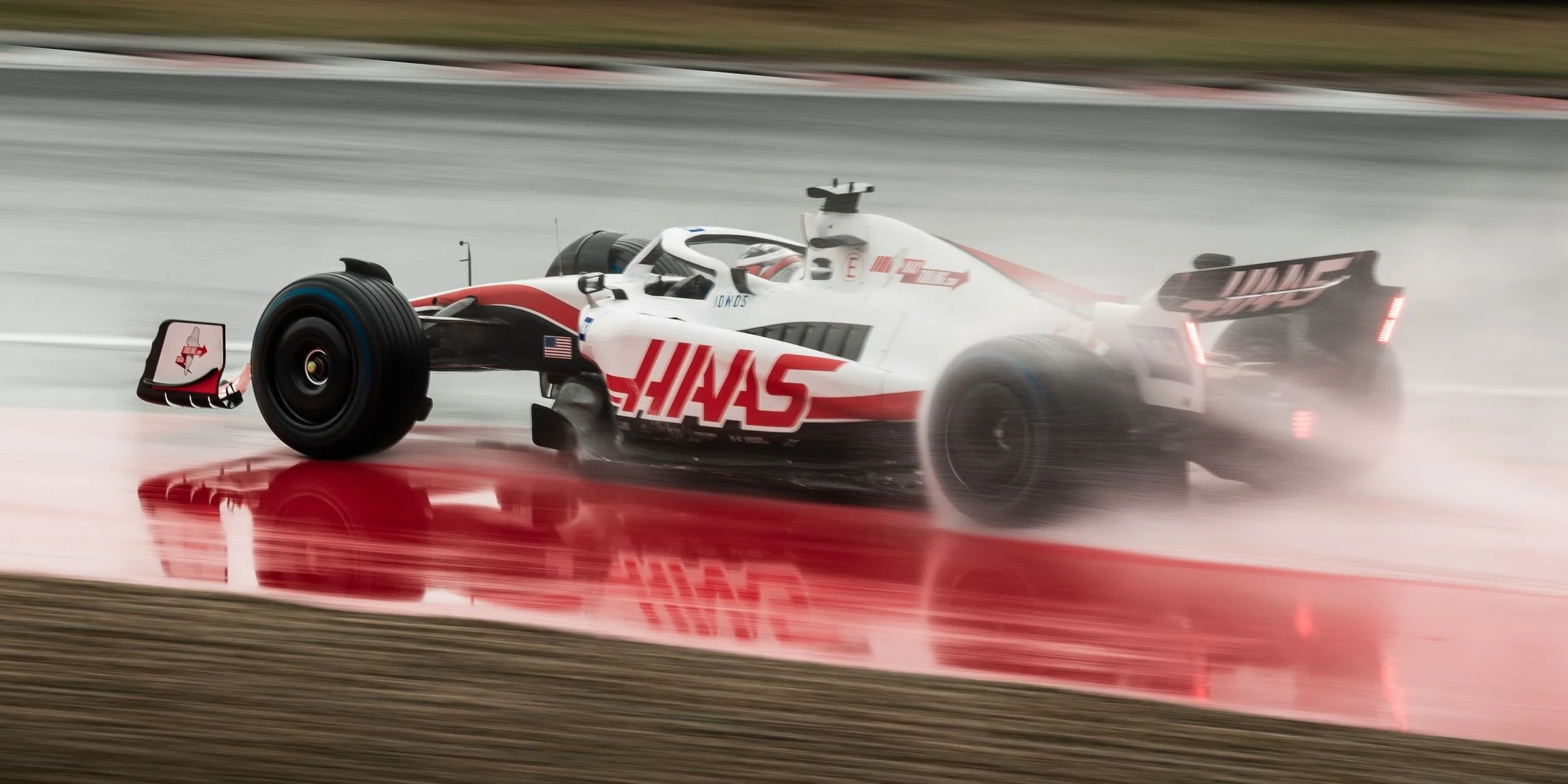 A racing car speeding through a wet race track, creating water spray behind it, with the Haas F1 Team logo on its side.