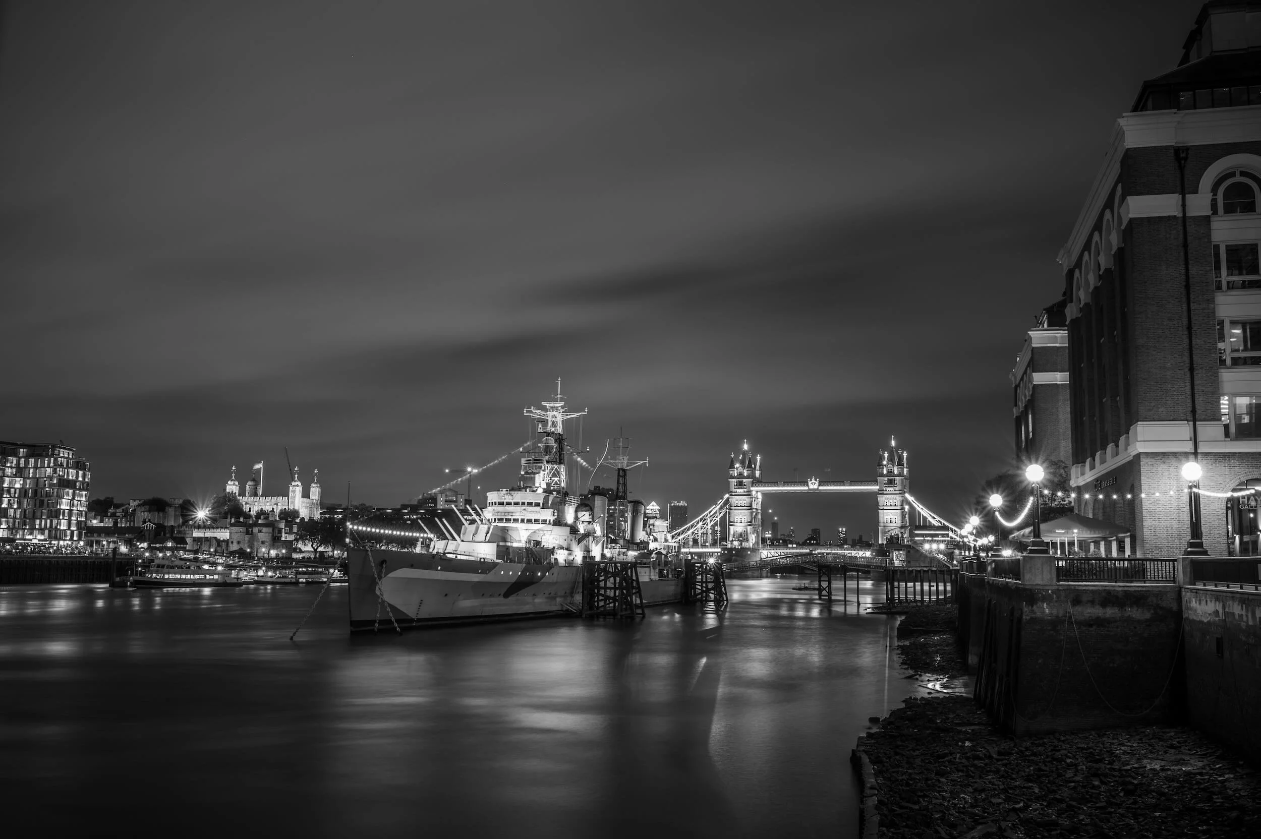 Night view of the Tower Bridge and the River Thames in London, with boats docked along the river and illuminated buildings in the background.