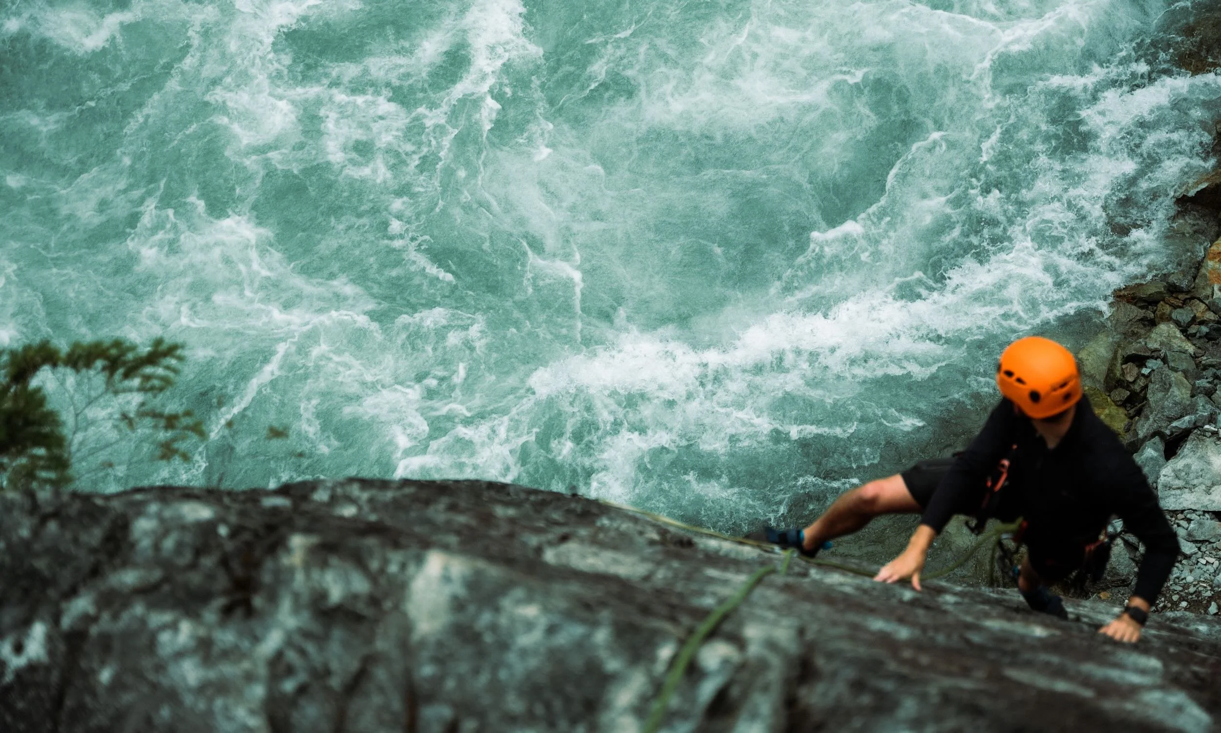 A person wearing an orange helmet and black clothing climbing a wet rocky ledge near a body of turbulent water.