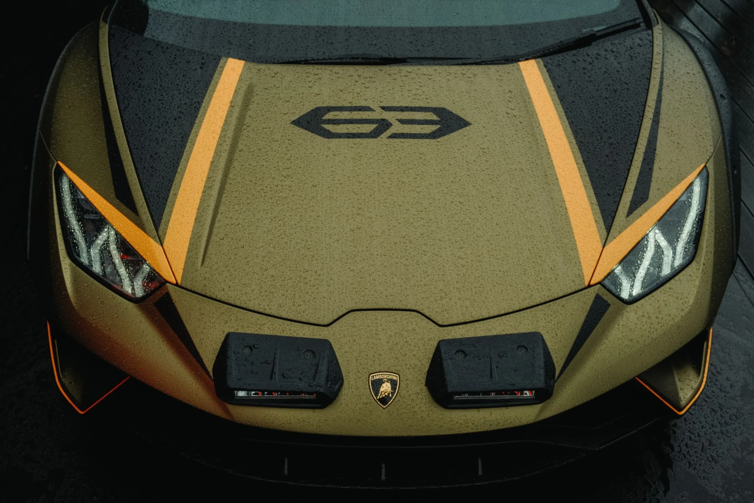 Front view of a gold Lamborghini sports car with black accents, a Lamborghini logo on the hood, and rain droplets on the surface.
