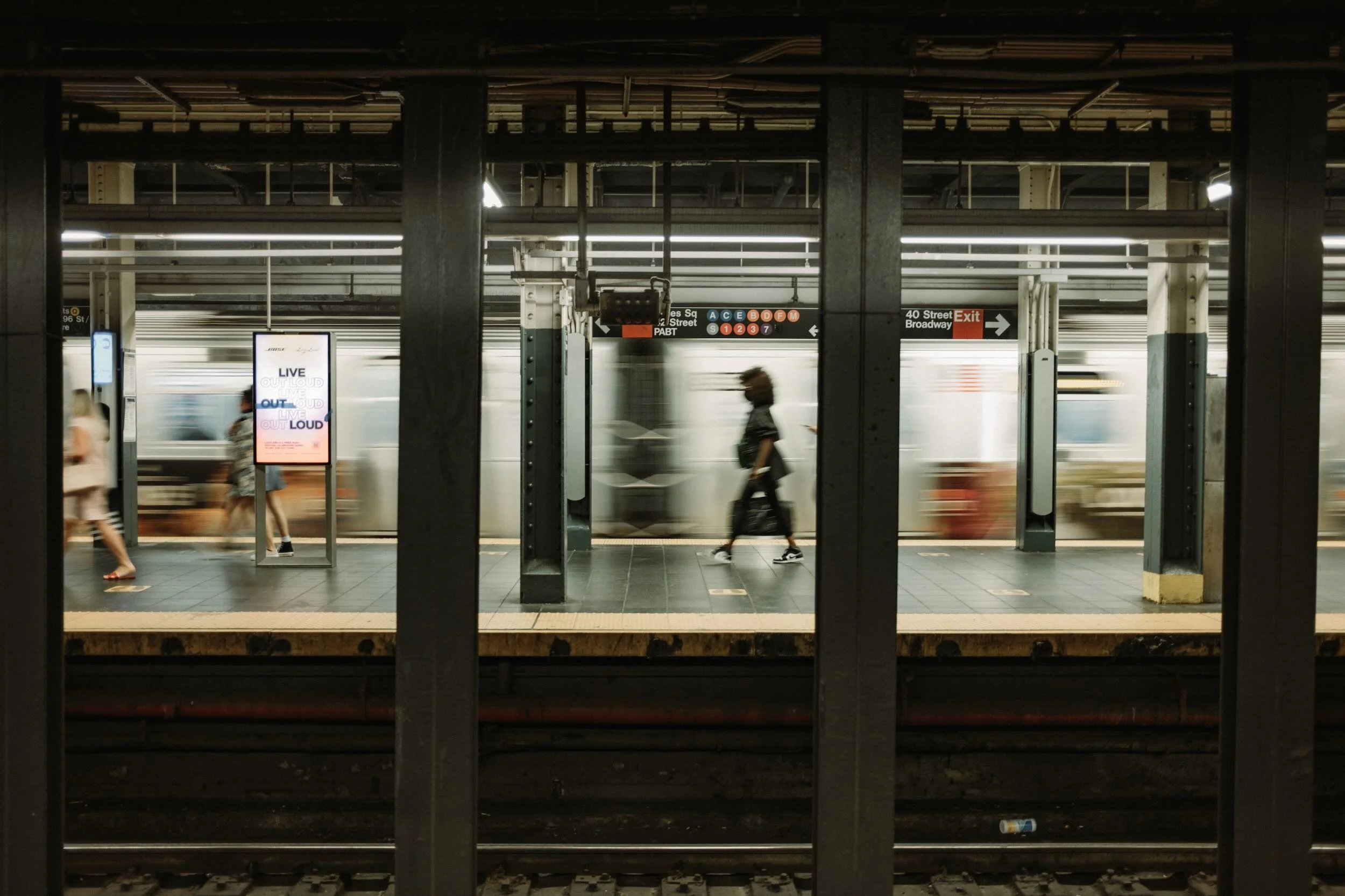 A subway platform with a moving train arriving, people walking across the platform, and signs indicating subway lines and an exit to 40th Street Broadway.