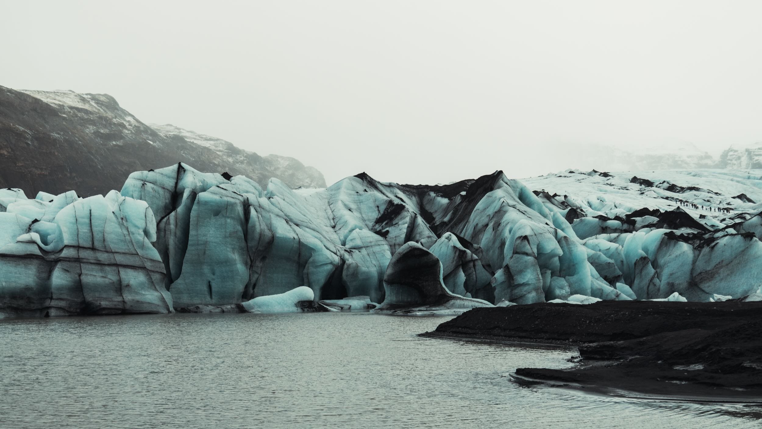A glacier with blue ice formations by a body of water, with mountains and an overcast sky in the background.