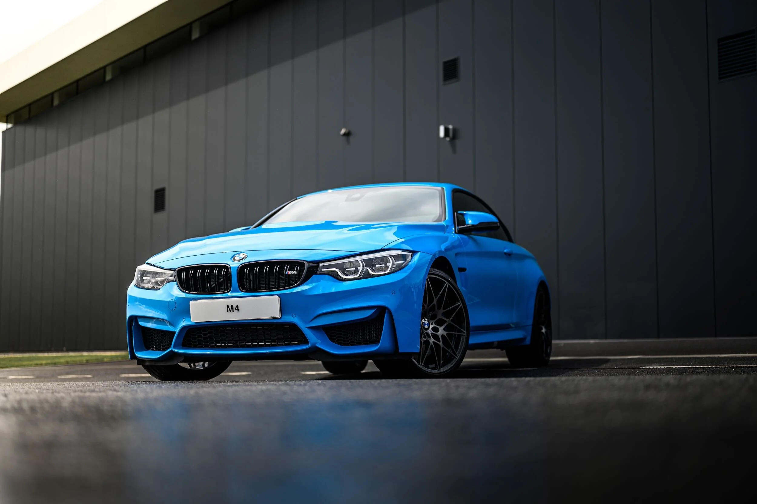 A bright blue BMW M4 sports car parked in front of a modern, gray building with clean lines and small windows.