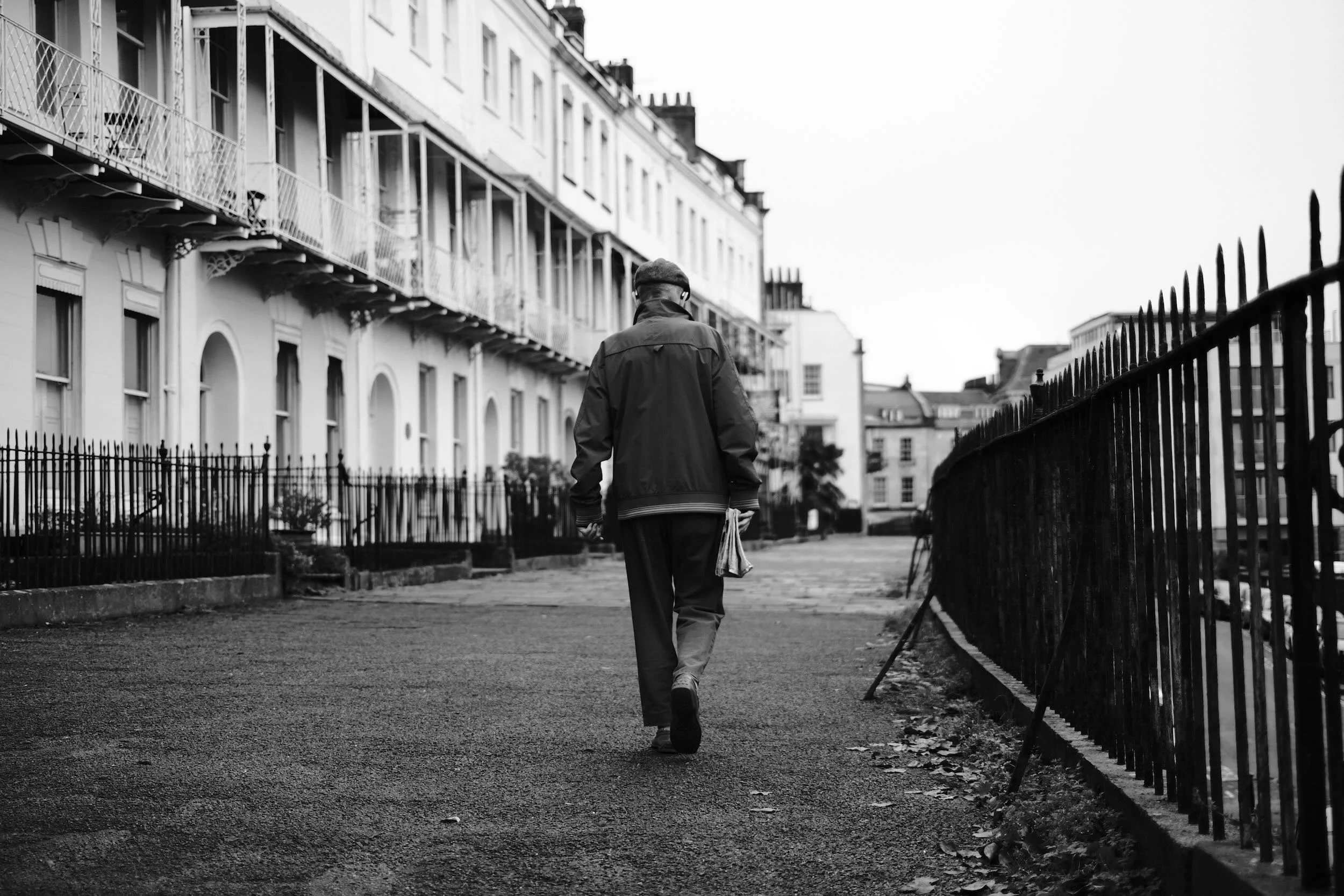A man walking along a city sidewalk in black and white, viewed from behind. He wears a hat and a coat, carrying a bag, with buildings and an iron fence along the street.