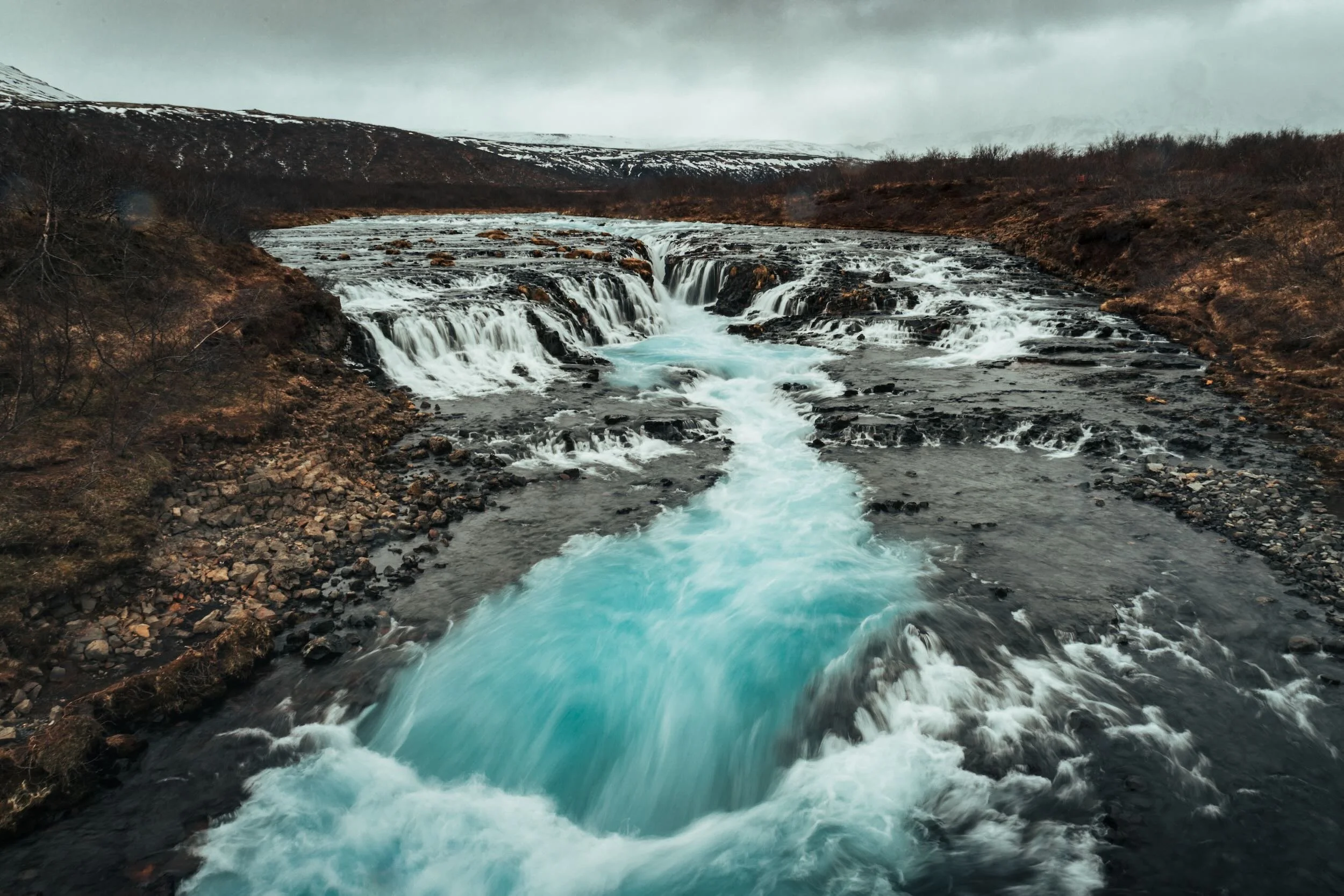 A river flowing through a rocky landscape in a cold, mountainous area with snow on the distant hills under a cloudy sky.
