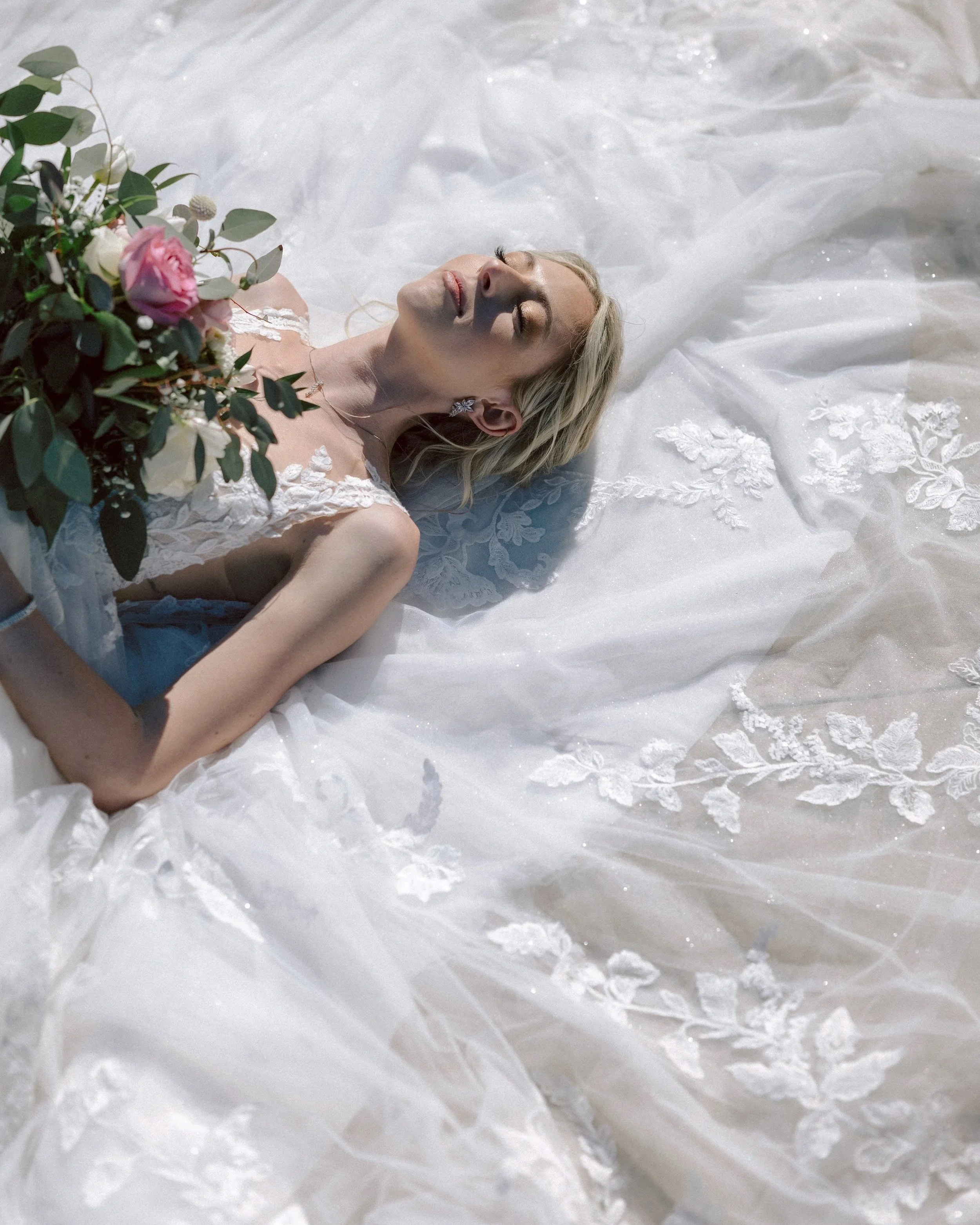 A woman in a wedding dress lying on the ground with her eyes closed, holding a bouquet of flowers.