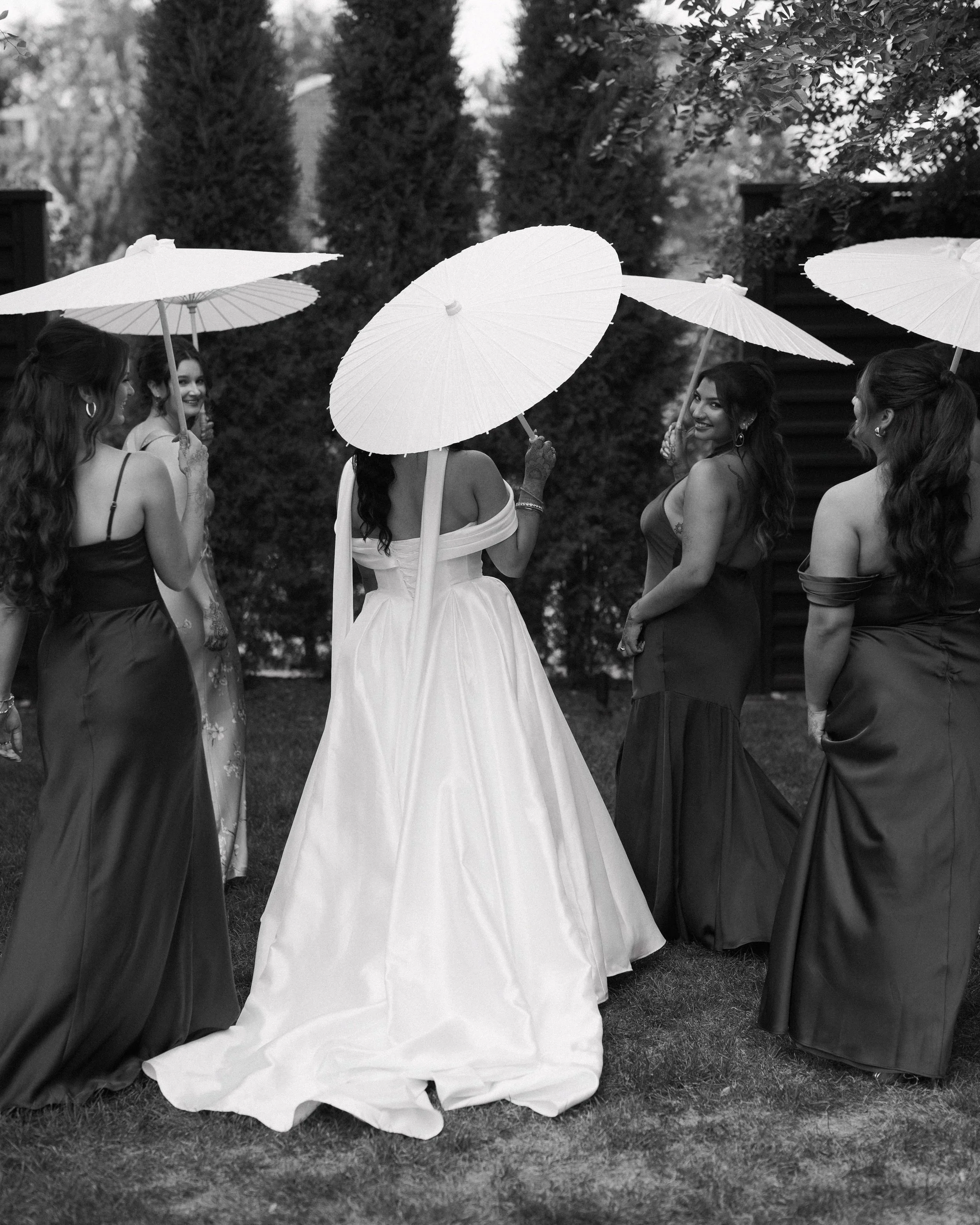 A group of women in formal dresses holding umbrellas outdoors, with trees in the background.