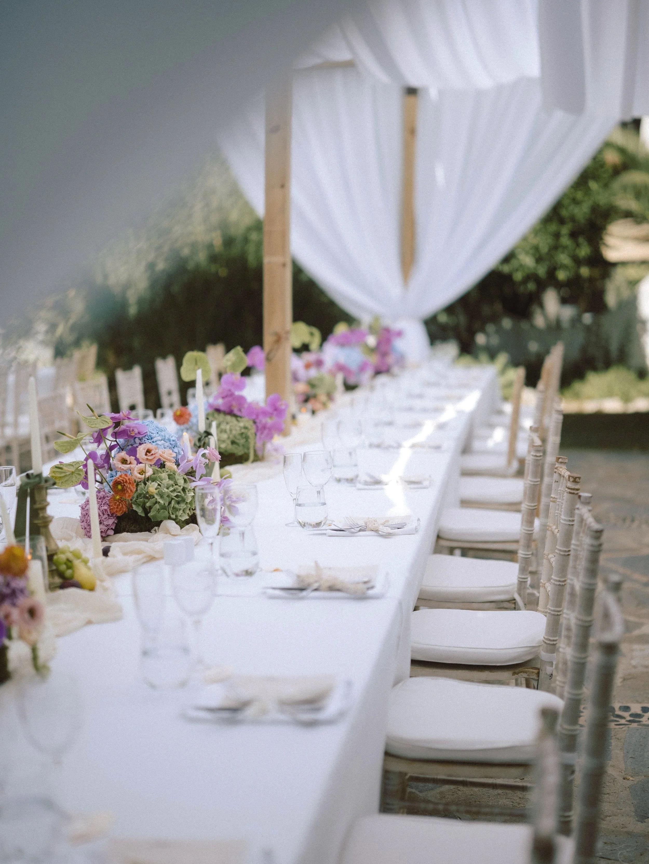 Long outdoor dining table decorated with colorful flowers, glassware, and white napkins, set under a white canopy with a garden background.