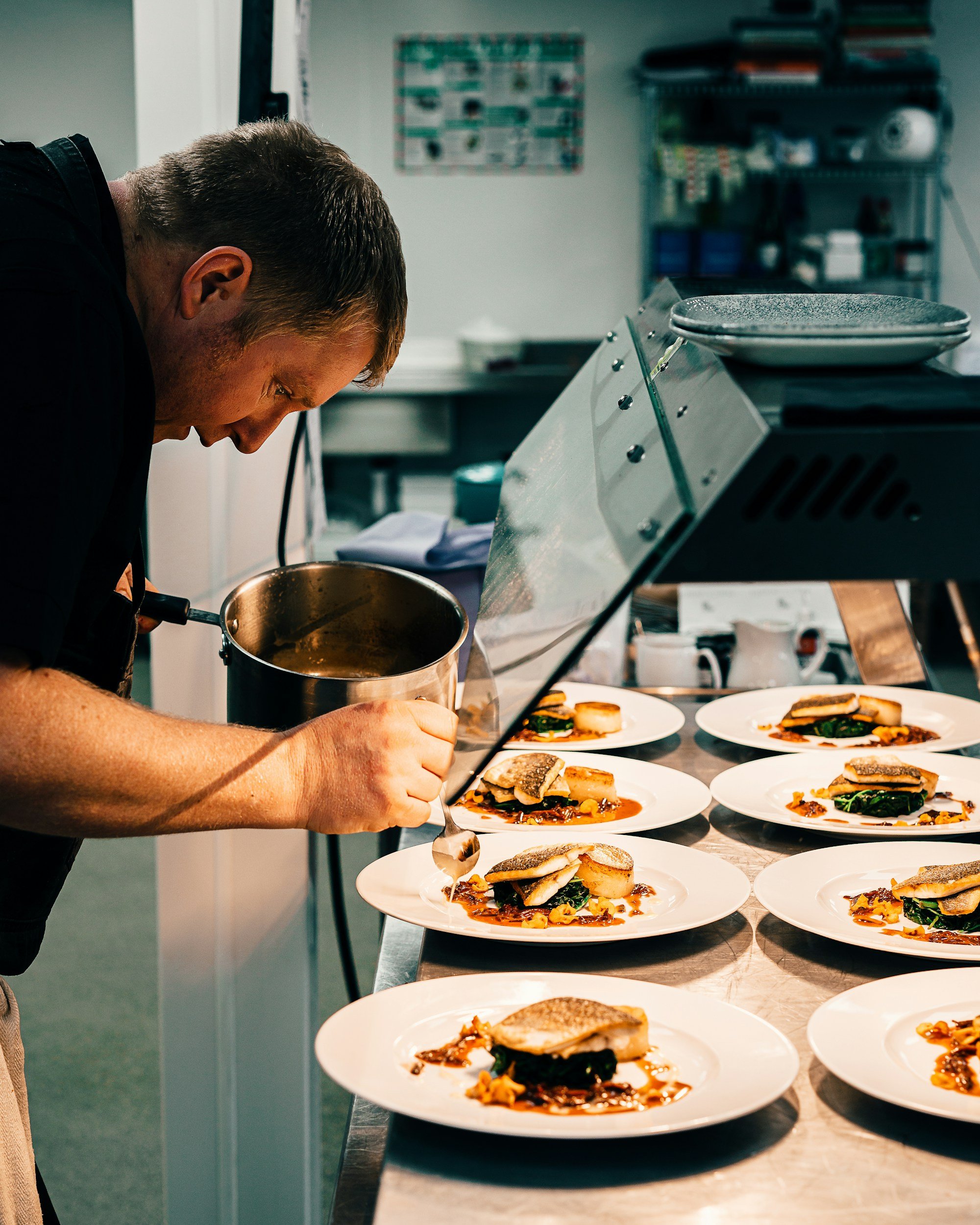 Un chef cuisinier au passe d'une cuisine professionnelle versant une sauce sur plusieurs assiettes de poisson dressées avec soin.