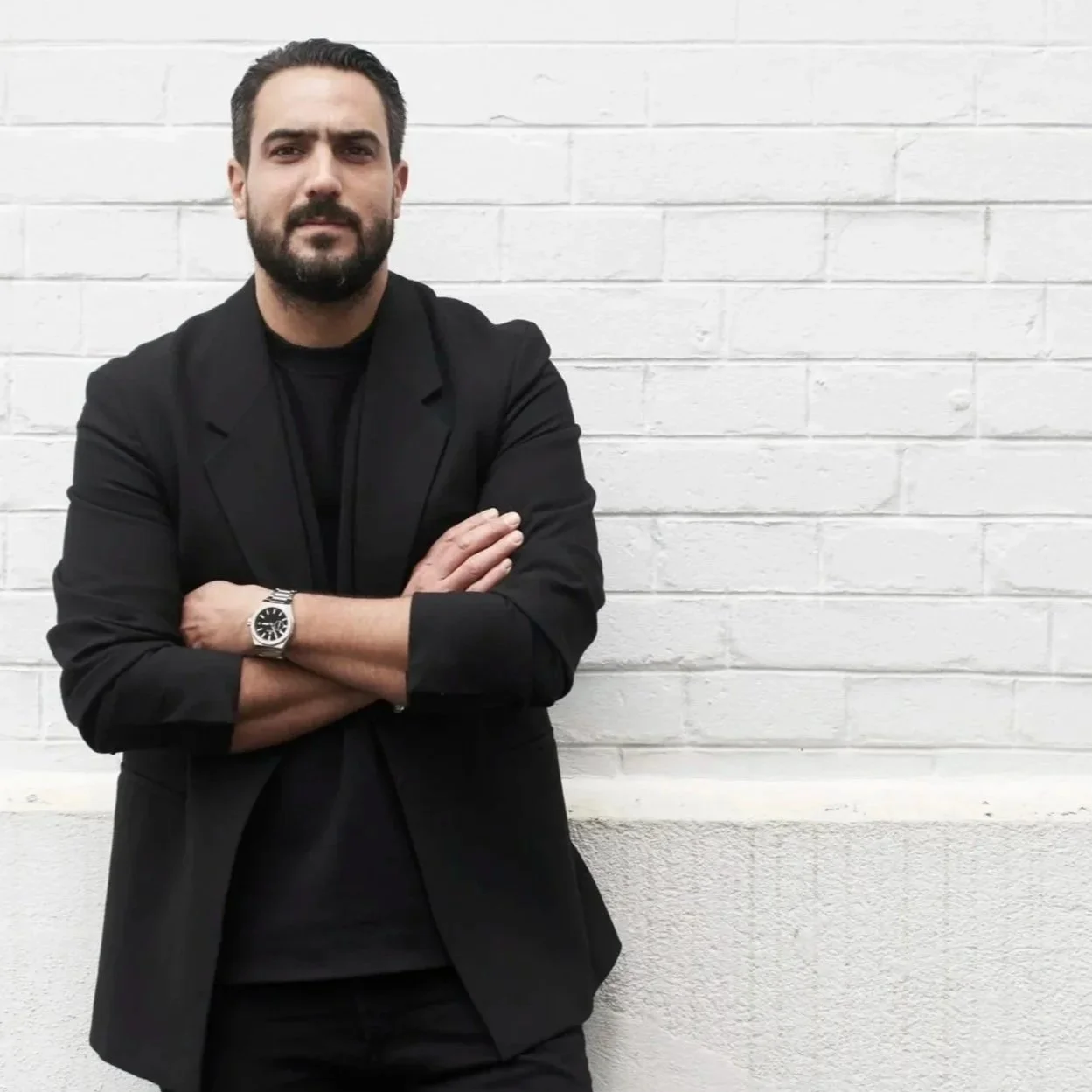Portrait du chef Jeffrey Cagnes vêtu d'une veste noire et arborant une montre de luxe Zénith devant un mur de briques blanches.