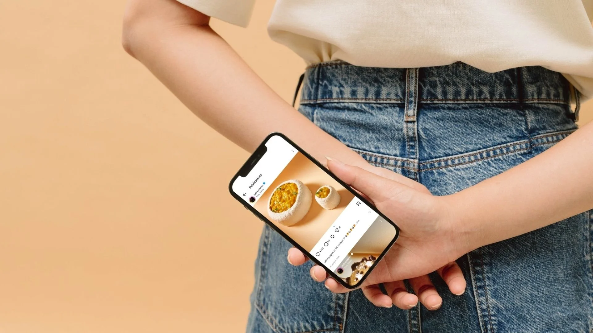 Gros plan sur une personne tenant un smartphone dans son dos, affichant une publication Instagram d'un plat culinaire sur un fond beige épuré.