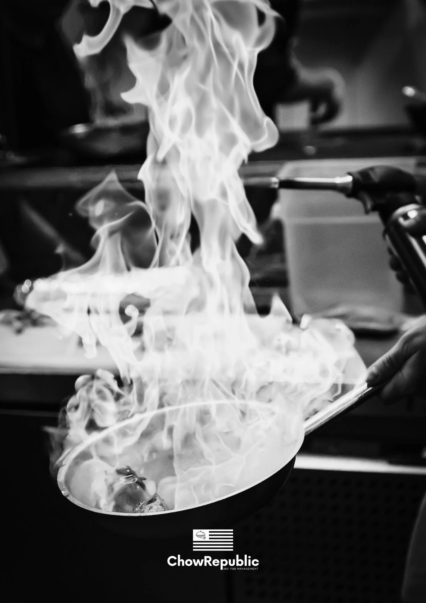 Image en noir et blanc de l'univers de la restauration montrant un coup de chalumeau et une poêle en plein flambage avec de grandes flammes, signée du logo Chow Republic.