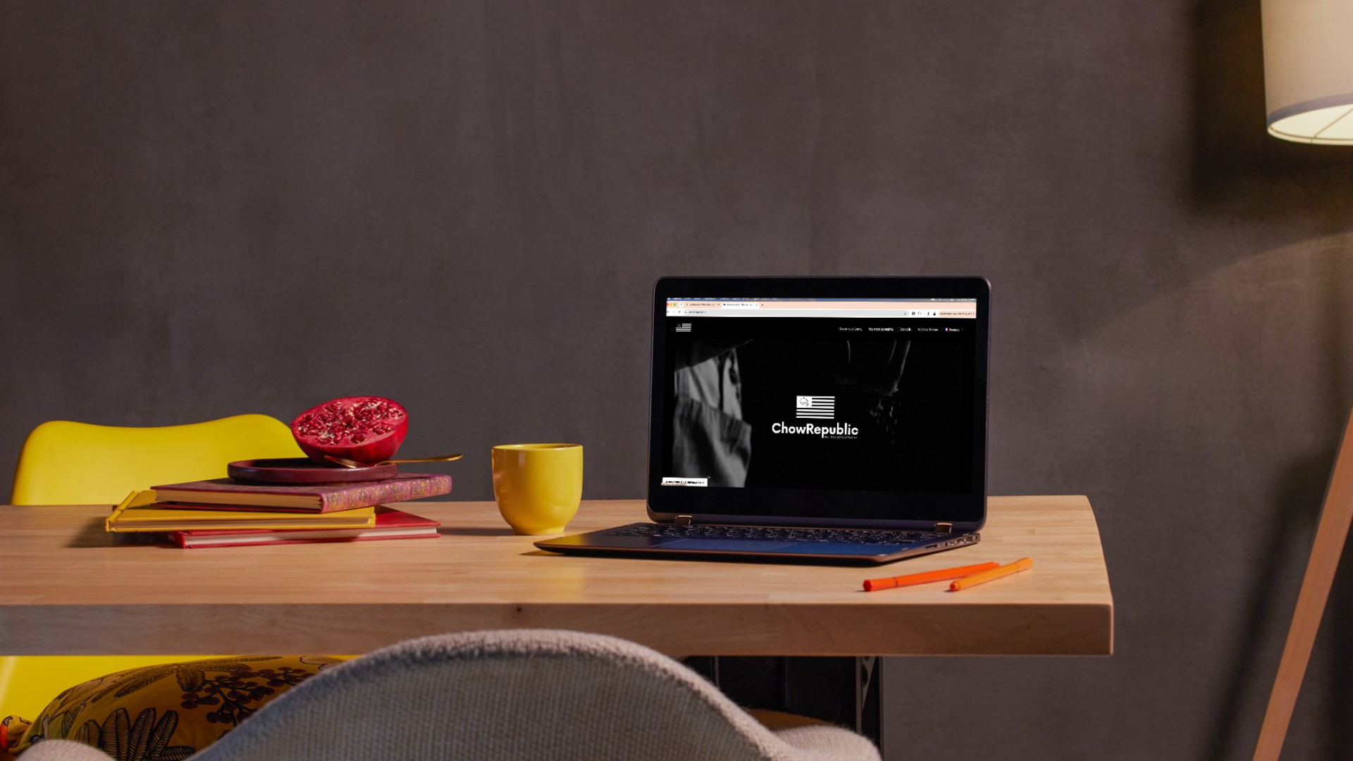 Un ordinateur portable ouvert sur le site de ChowRepublic, posé sur une table en bois à côté d'une tasse jaune, de livres et d'une grenade coupée en deux.