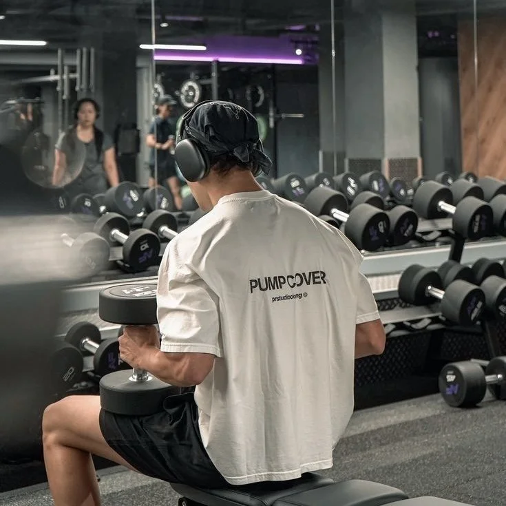 Person in a gym sitting on a bench, wearing headphones and a black cap, lifting a dumbbell with their right arm. The person has on a white t-shirt with the text "PUMP COVER" on the back, and is reflected in mirrors showing more gym equipment and other people exercising.