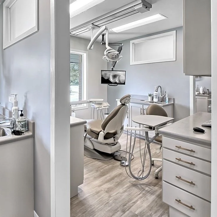 A dental examination room with a dental chair, overhead light, monitors, and dental tools in a clean, modern setting.